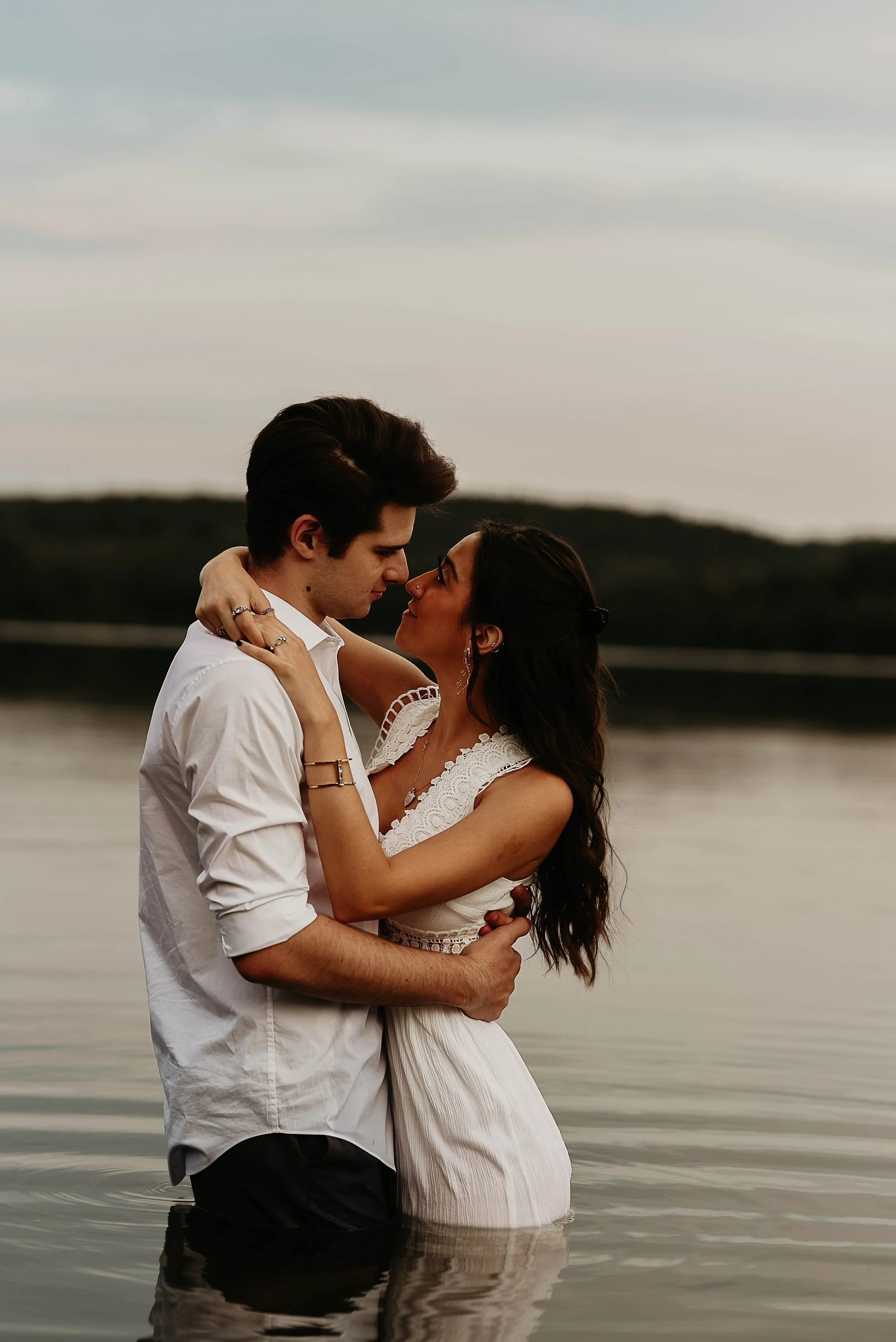 A couple stands in water, embracing and about to kiss, with a lake and distant hills in the background during sunset.