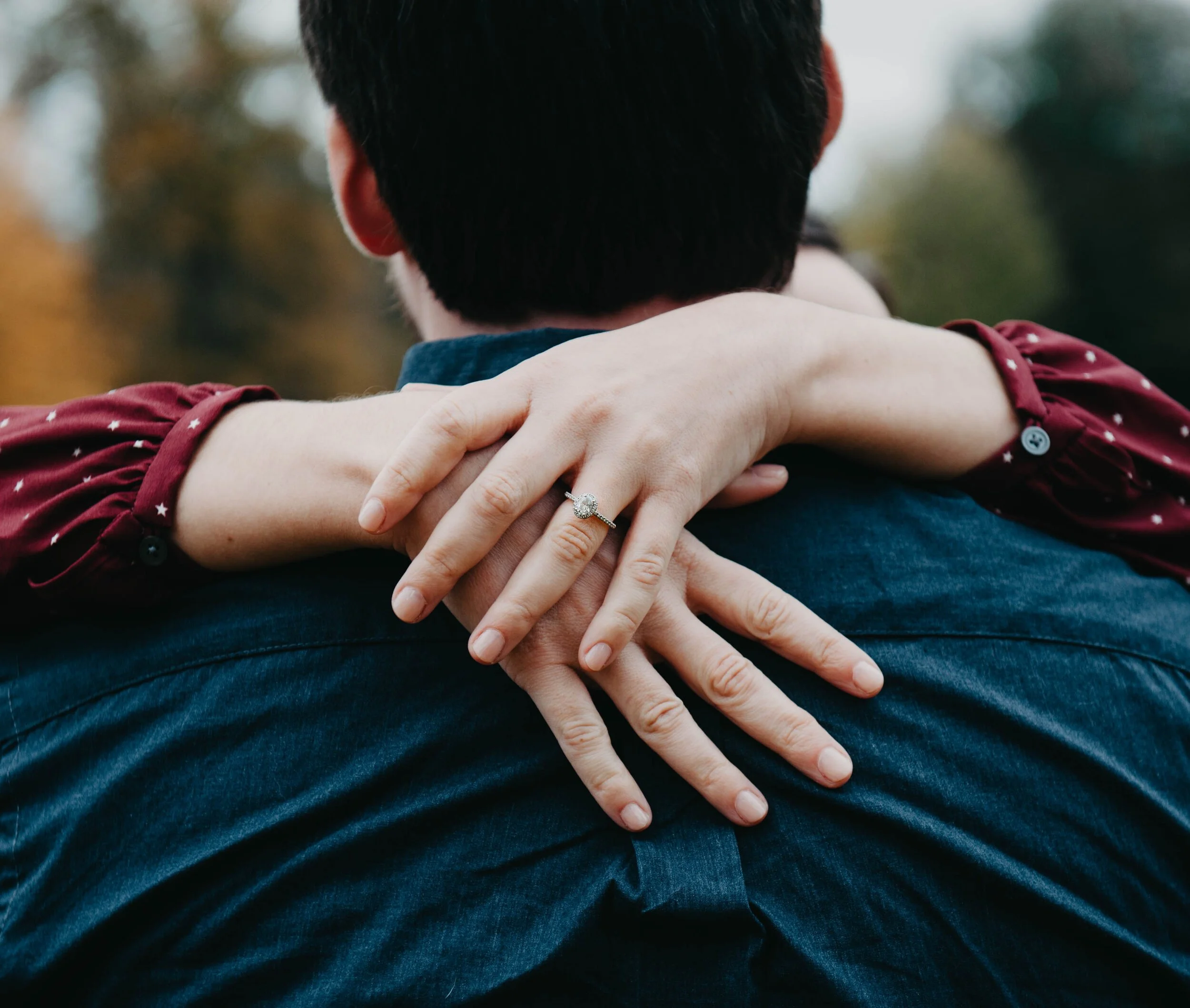 Close-up of a woman’s hands resting on a man’s shoulders, with her left hand showing an engagement ring. The man is facing away, wearing a denim jacket, with blurred outdoor background.
