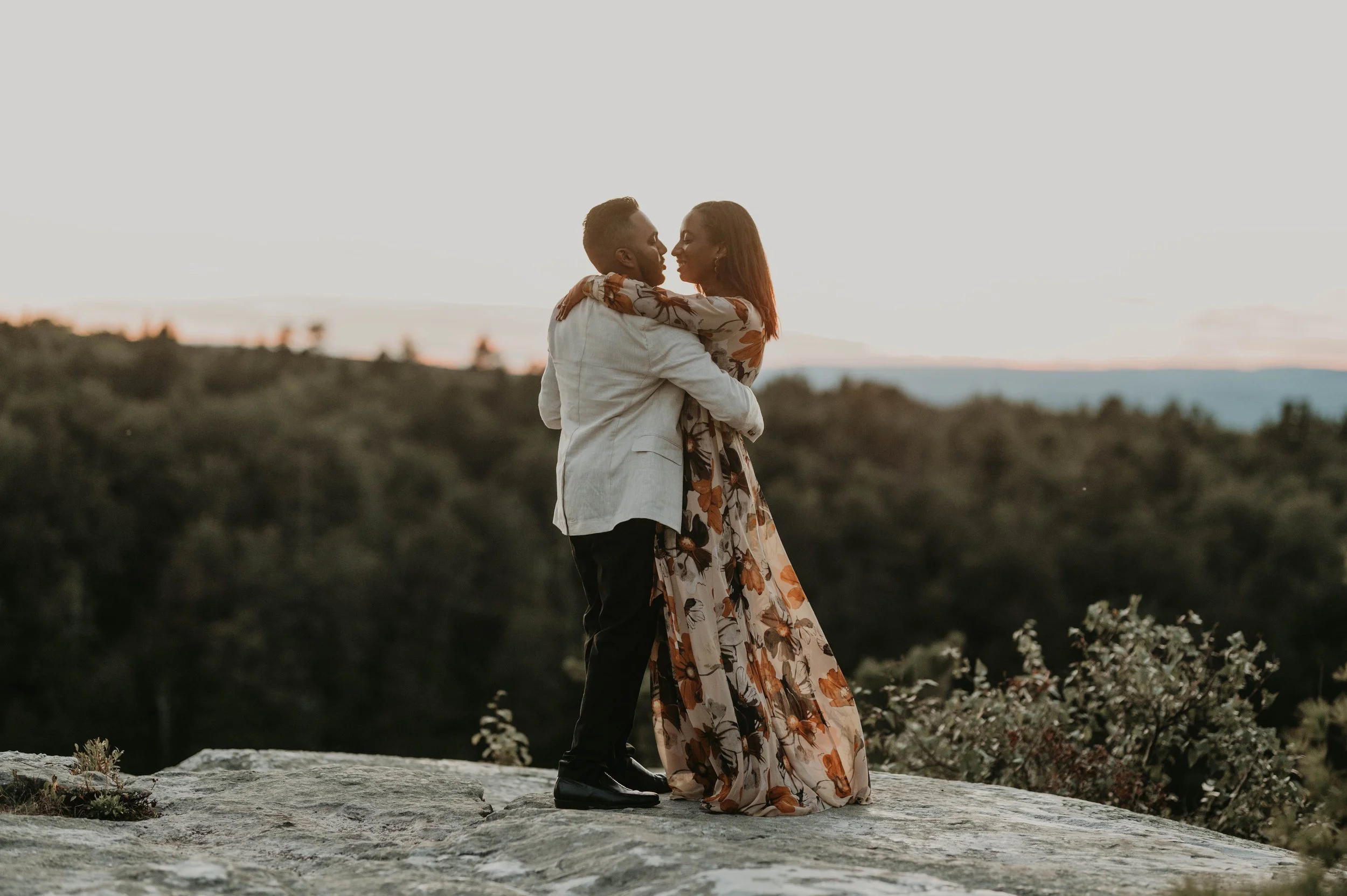 A couple embracing and about to kiss on a rock with a scenic landscape of trees and hills in the background during sunset.
