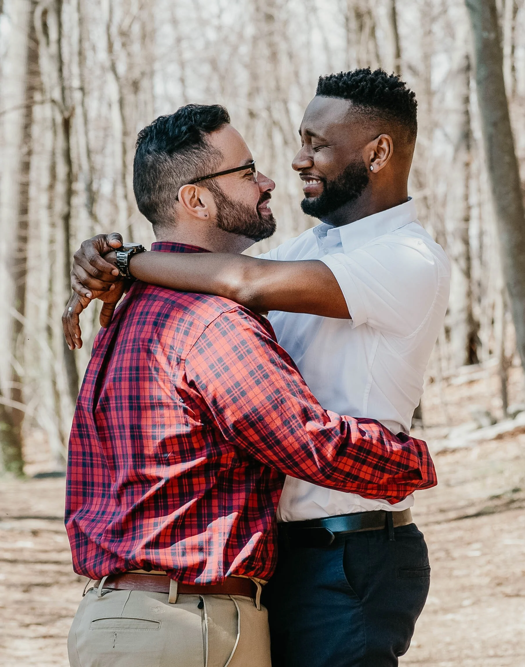 Two men embracing outdoors in a forest, looking at each other and smiling.