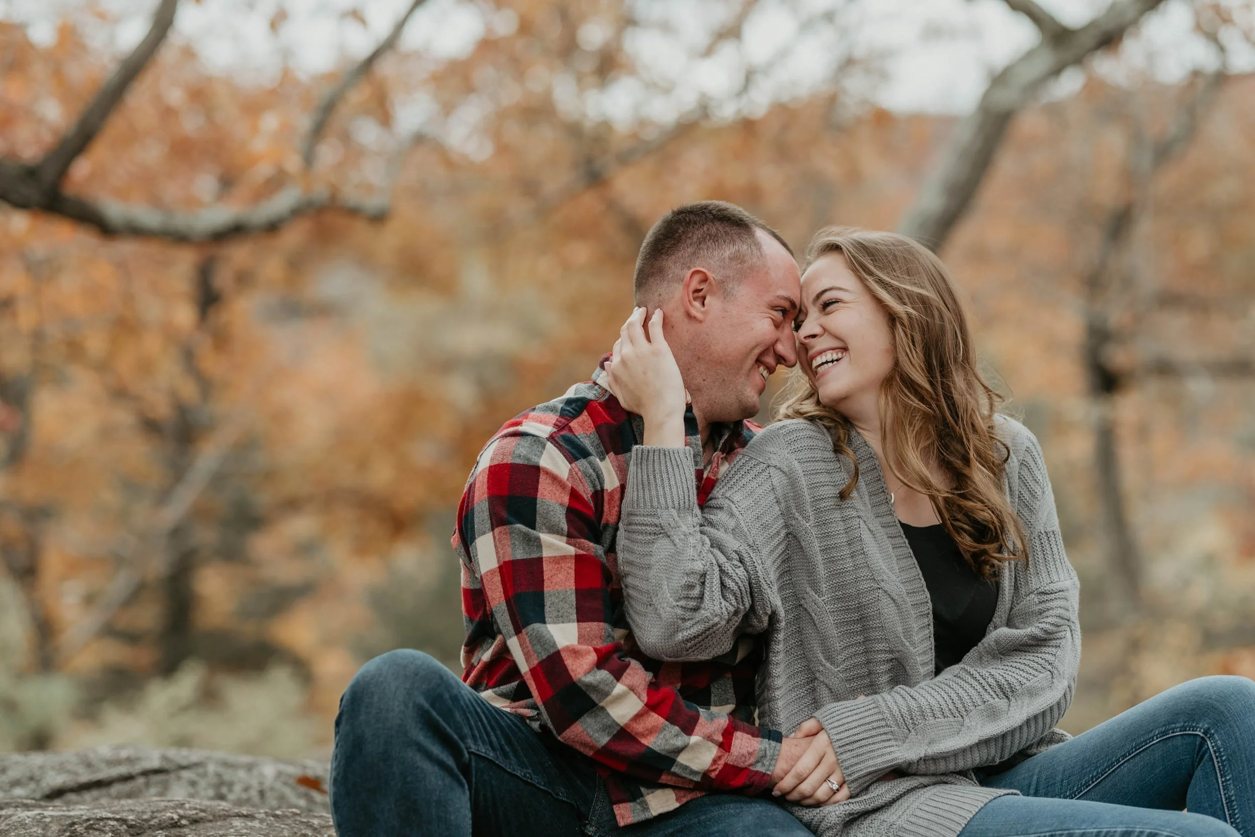 A couple sitting outdoors on a rock in autumn, smiling and touching foreheads with their eyes closed, surrounded by fall-colored trees.