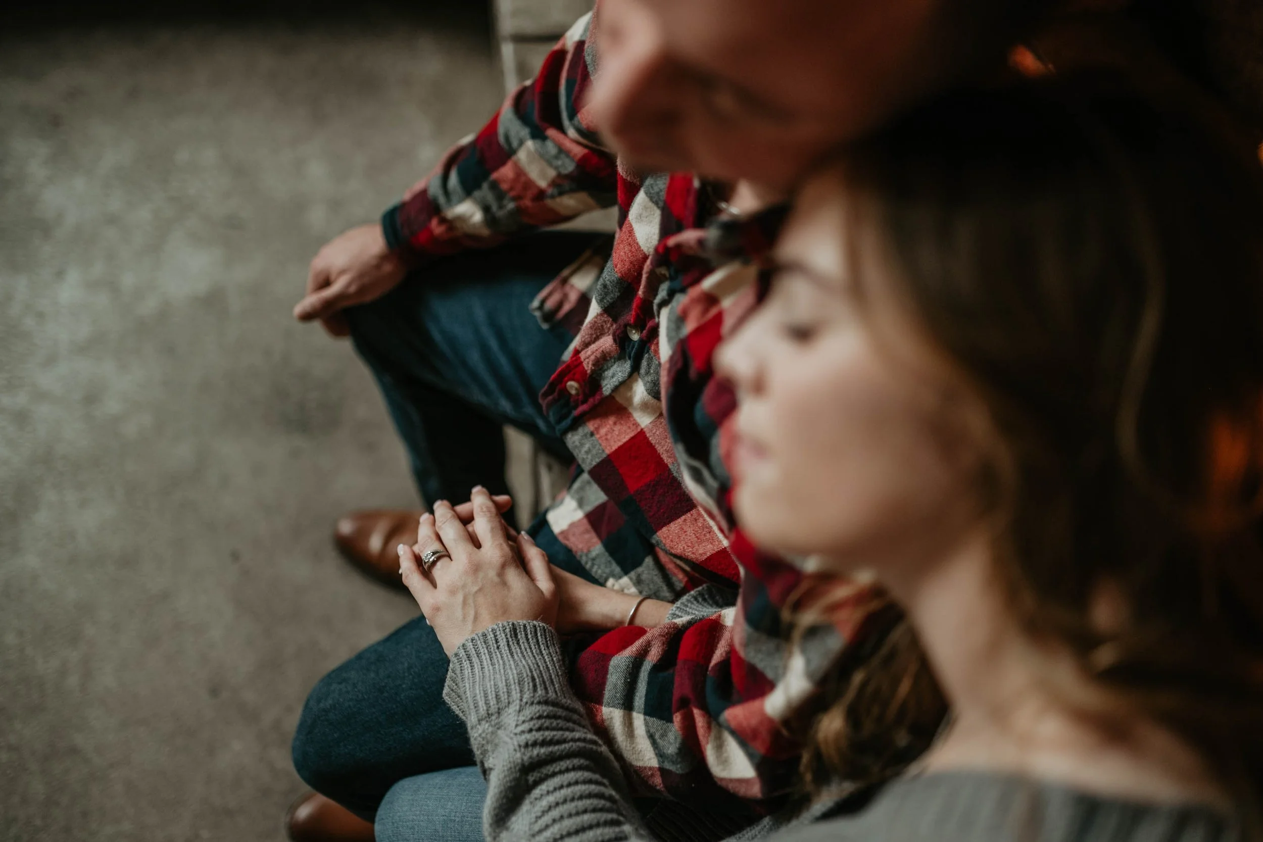 A man and woman sit closely together, the woman resting her head on the man's shoulder, holding hands. They are wearing casual clothing, and the photo is taken from above.