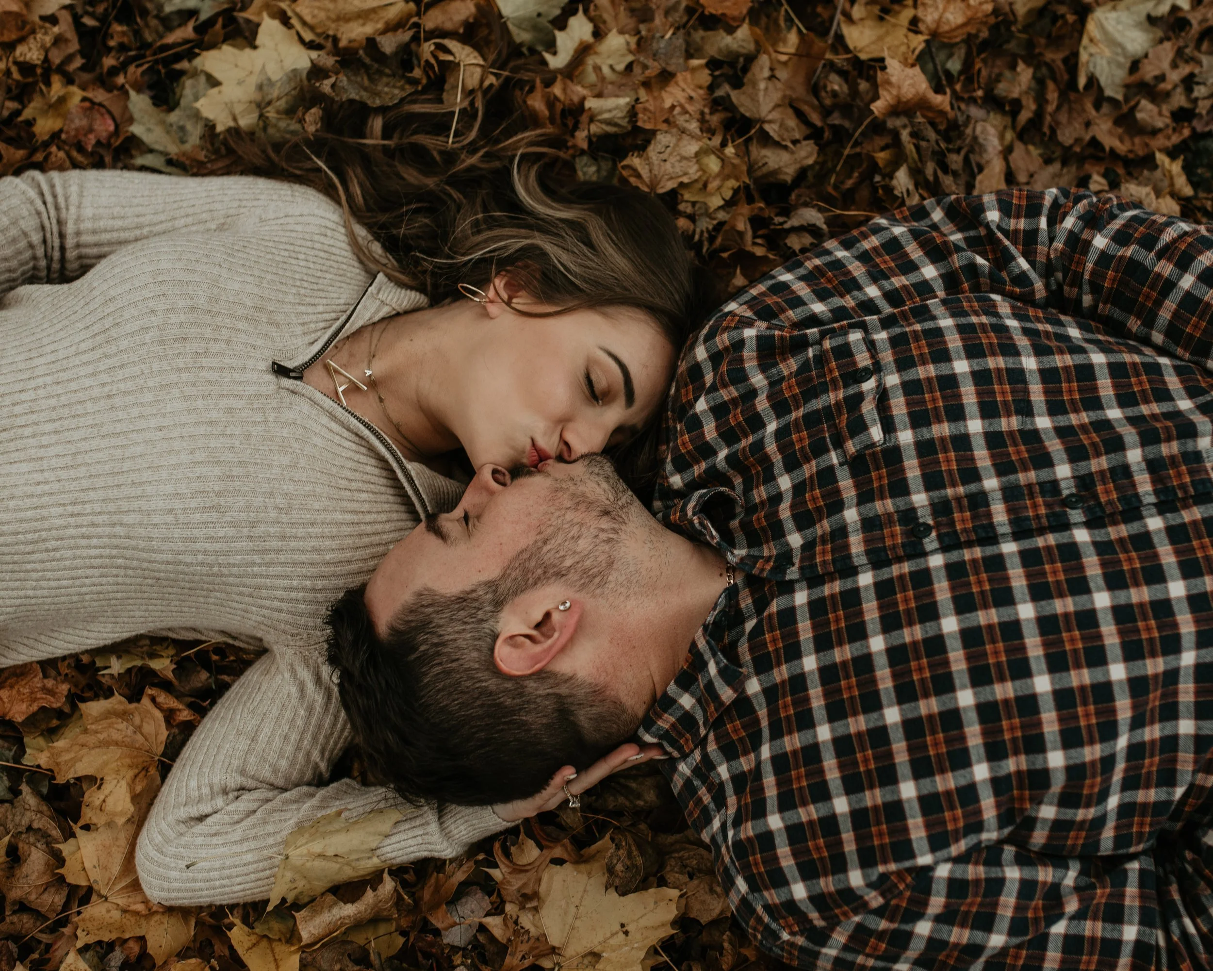 A young couple lying on the ground covered with fallen leaves, facing each other and touching foreheads, sharing a kiss.
