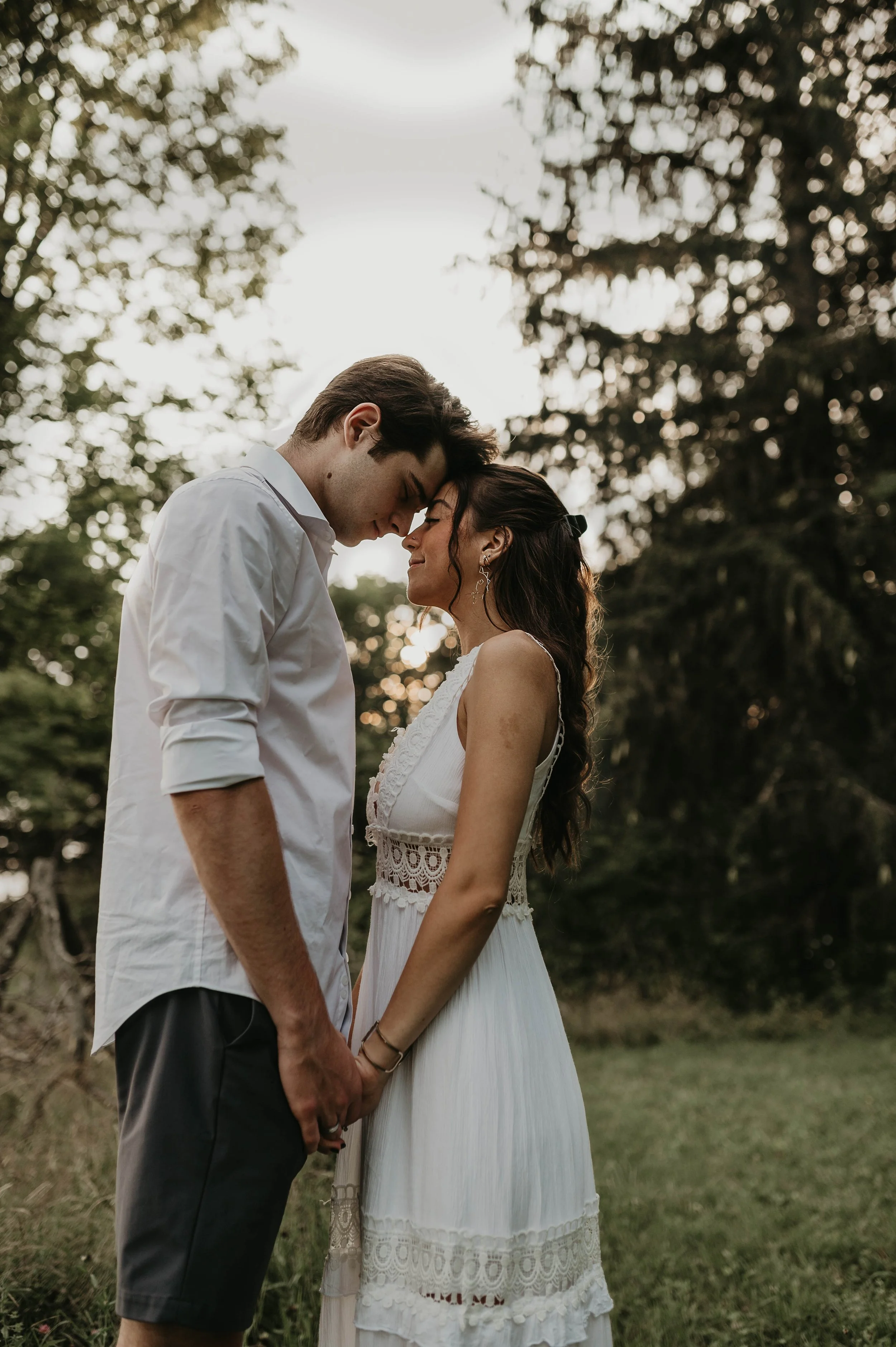 A young couple, a man and a woman, are standing close together outdoors, touching foreheads and holding hands. The man is wearing a white shirt and dark shorts, while the woman is wearing a white sleeveless dress with lace details. They are surrounde