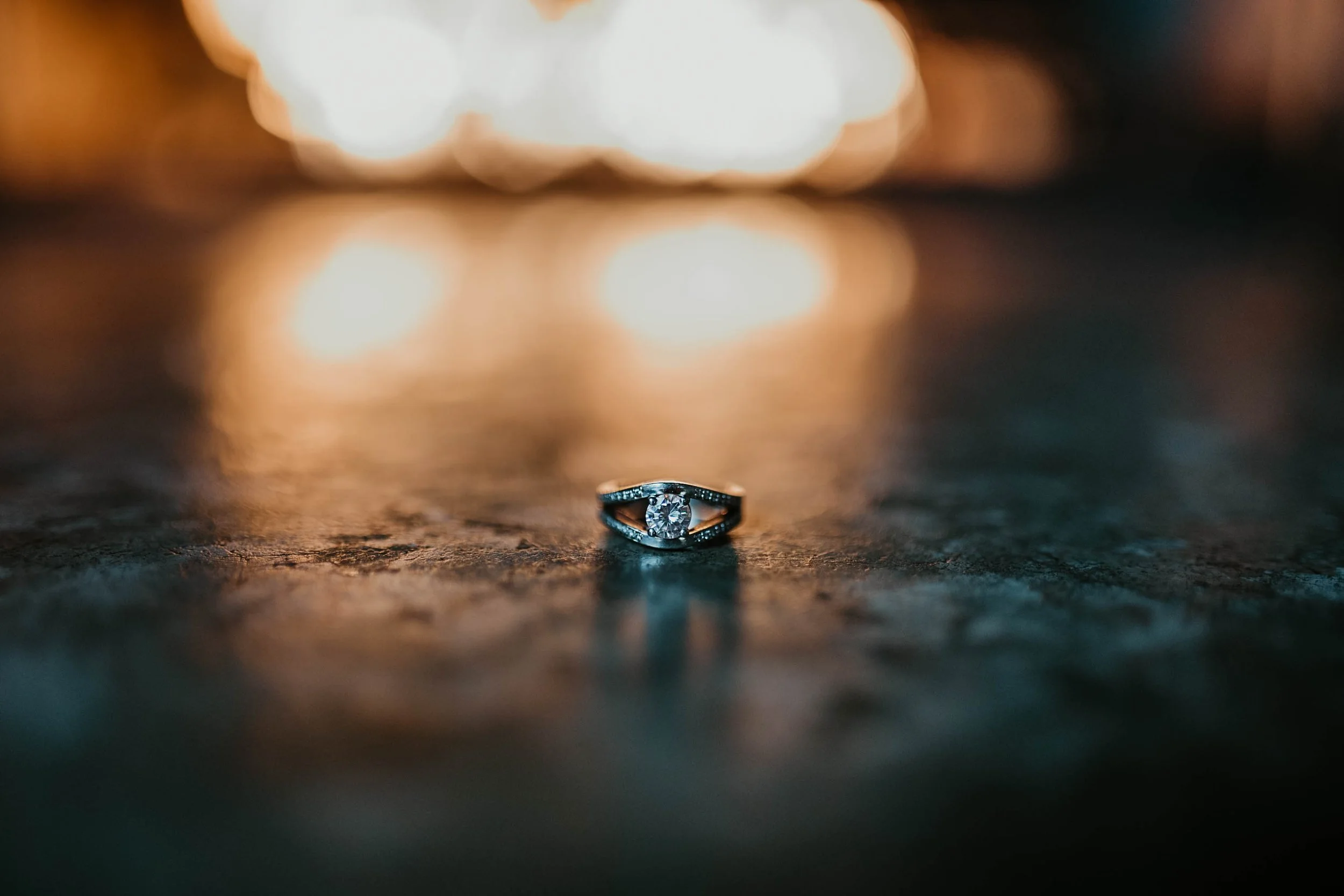 A diamond engagement ring resting on a dark textured surface with blurry warm lights in the background.