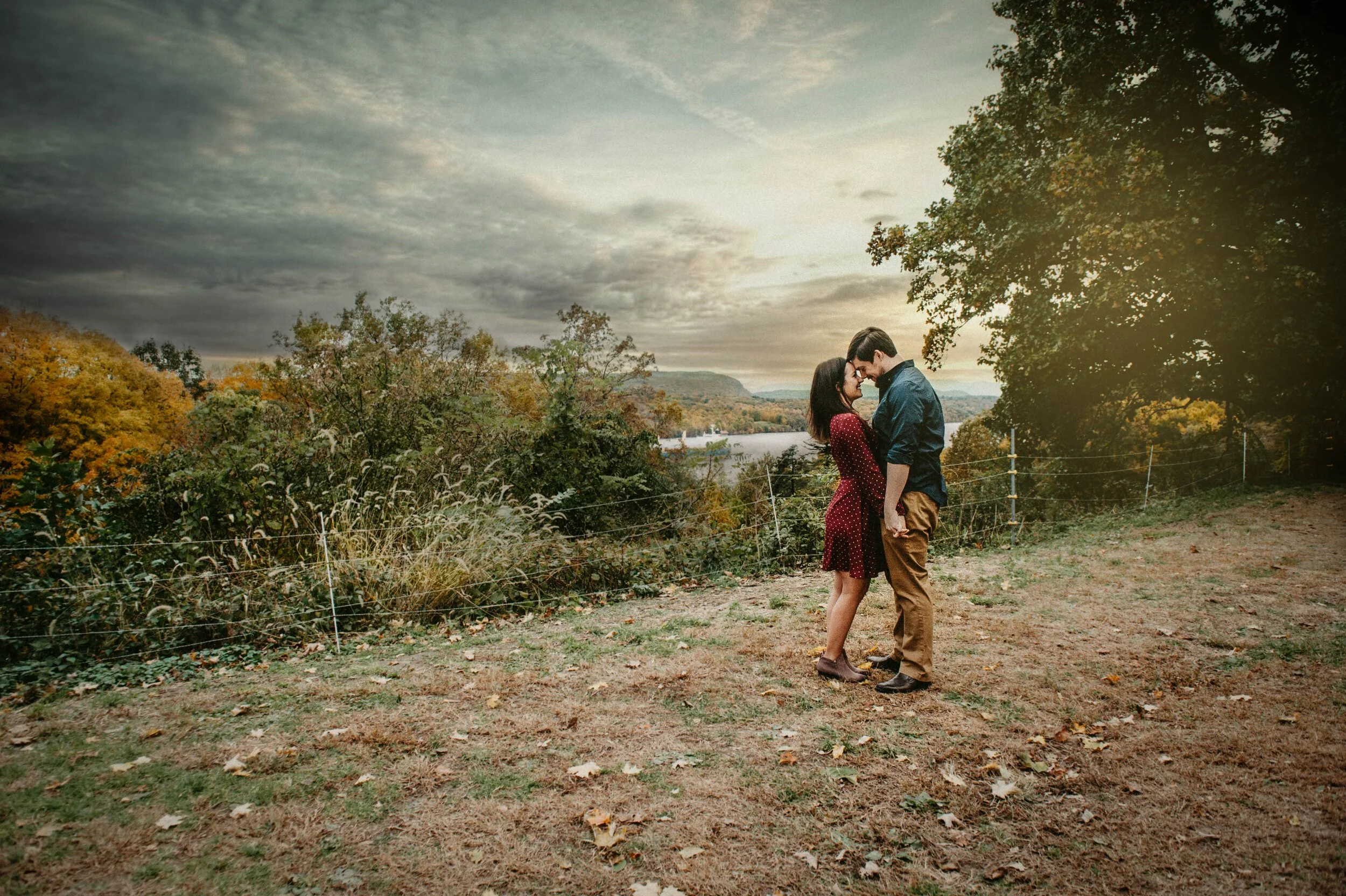 A couple stands close together outdoors on a hillside, holding hands and leaning their foreheads together. They are surrounded by autumn foliage under a cloudy sky, with a river and hills in the background.