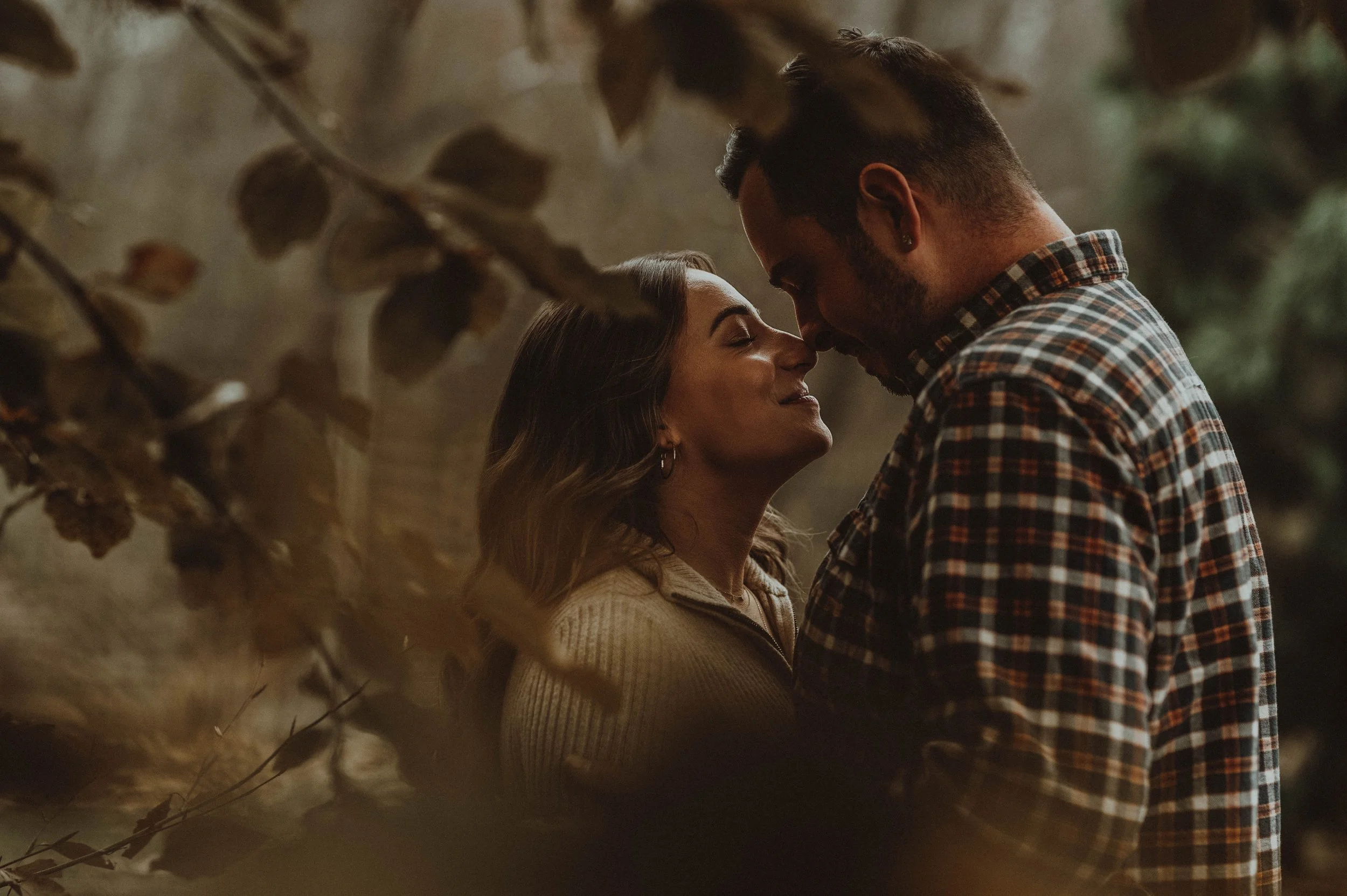 A couple sharing an intimate moment outdoors, with their faces close and eyes closed, surrounded by autumn leaves.