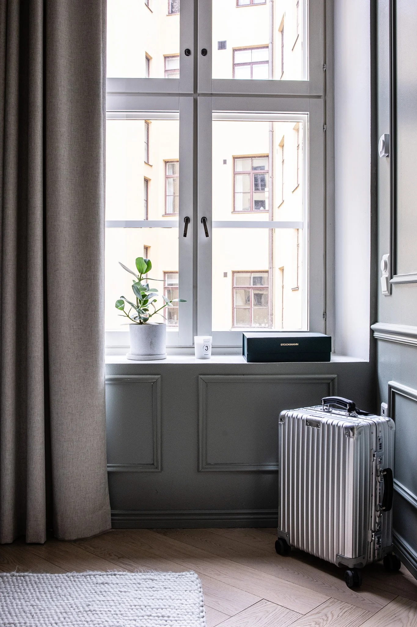 Ikkunan ääressä on vihreä kasvi ja pieni valkoinen kynttilä, tupakkalaatikko ja hopea matkalaukku. Taustalla näkyy rakennus ja taivasta.