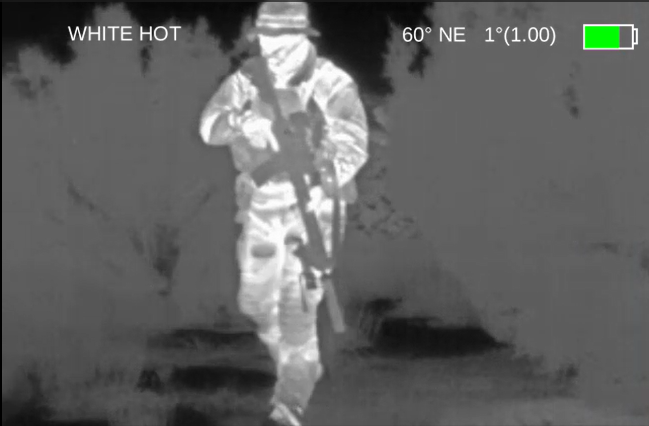 White hot thermal image of a subject wearing tactical equipment and carrying a scoped rifle among several trees.