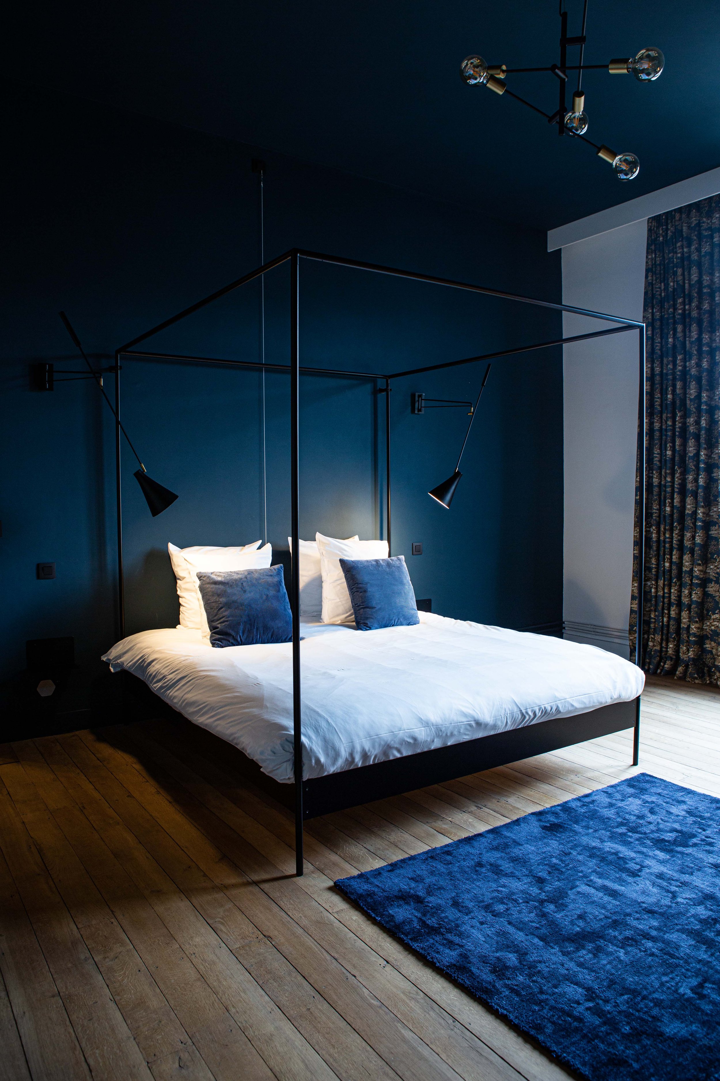  Une chambre au coloris bleu nuit dans laquelle on retrouve au lit à baldaquin moderne et des lampes design.  