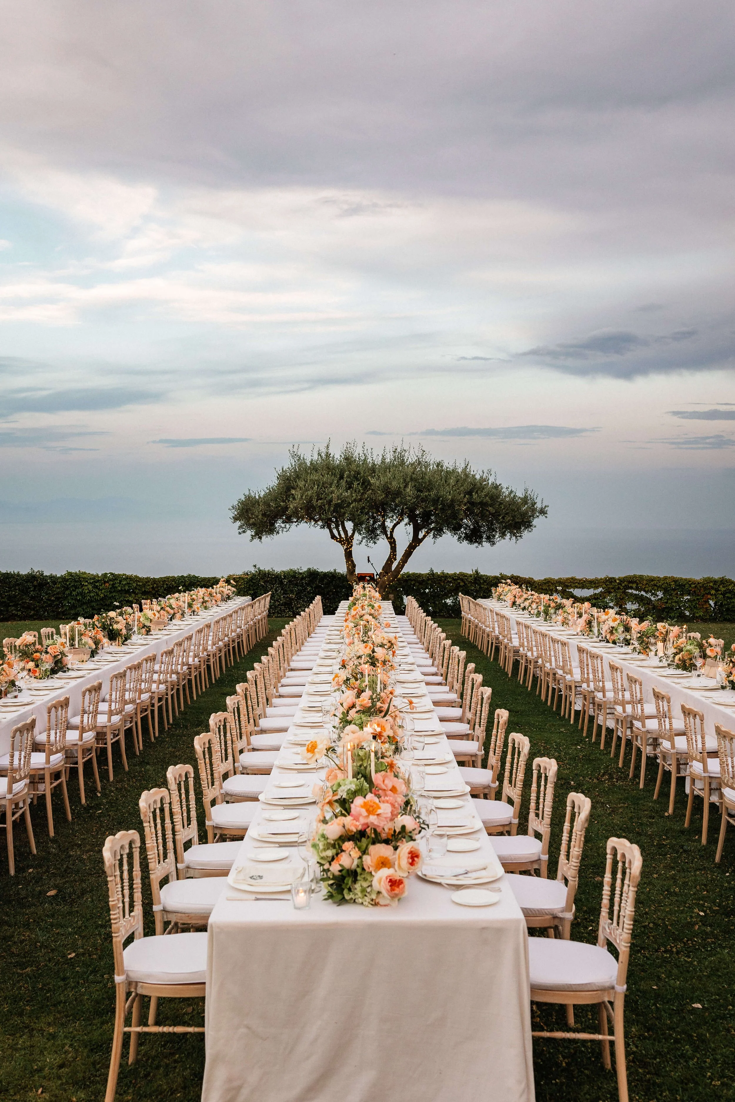 Outdoor wedding reception setup with long white-draped tables, decorated with peach and pink floral arrangements, surrounded by white chairs. A tree stands in the background under a cloudy sky.