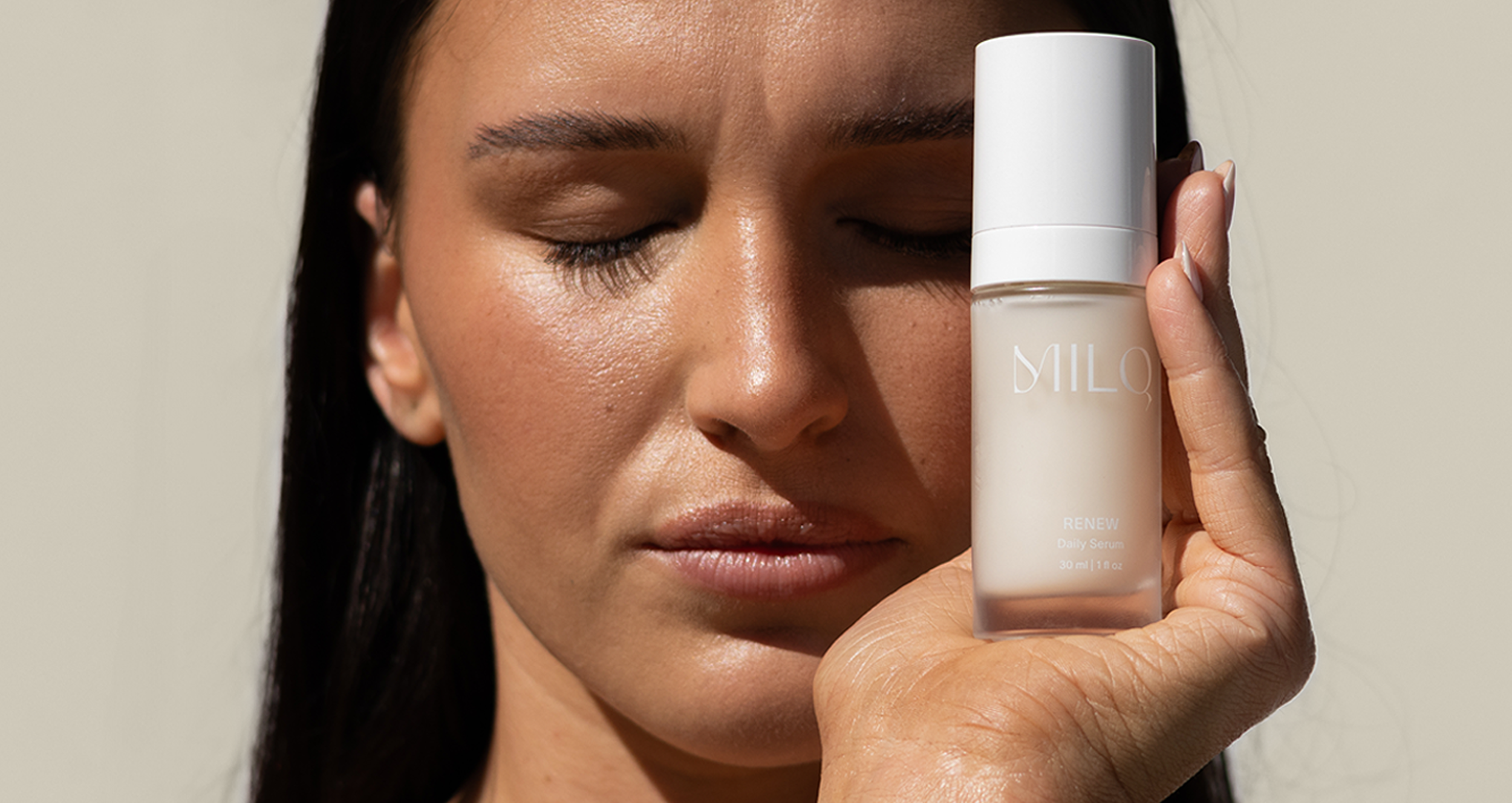 A woman with closed eyes holding a white bottle labeled "MILQ Renew Daily Serum" close to her face, with sunlit skin and dark hair.