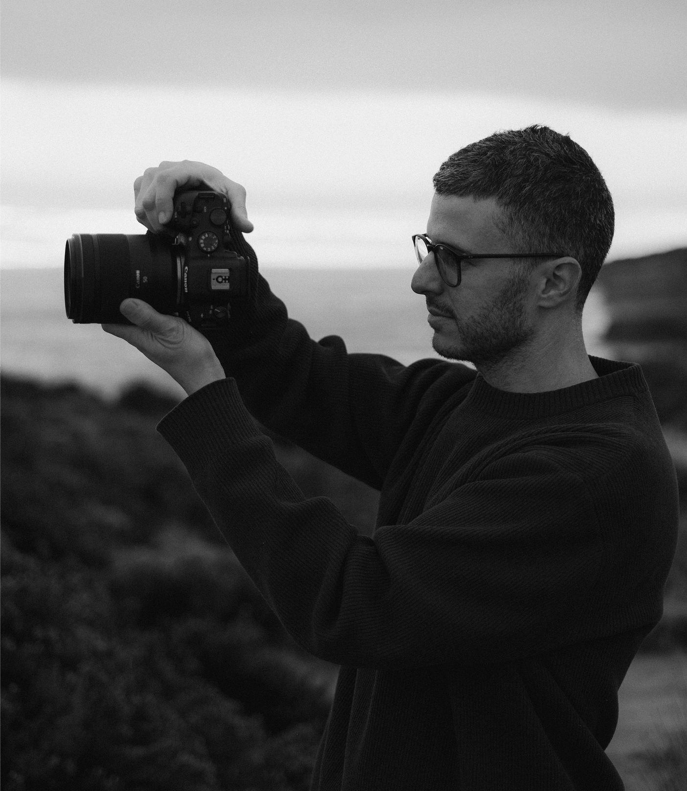 A man with glasses and short hair holding a camera outdoors during dusk or dawn, with a blurred landscape background.