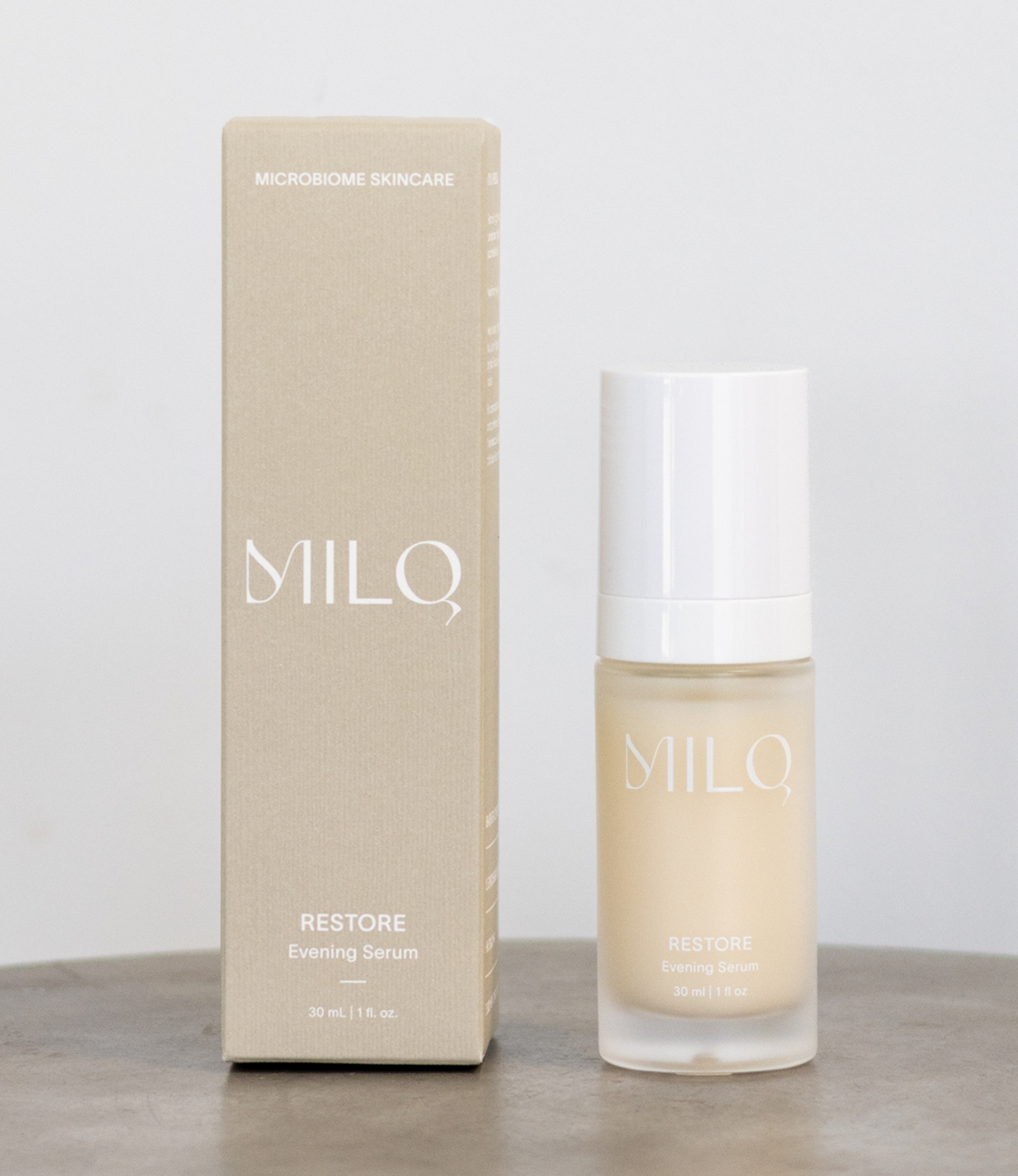 A beige box and a frosted glass bottle of MILQ Restore Evening Serum on a gray surface with a plain white background.