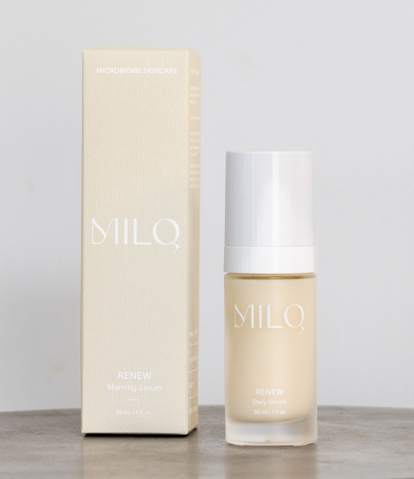 A skincare product set including a box and a bottle with a white cap, labeled 'MilQ RENEW Daily Serum' on a gray surface against a plain light background.