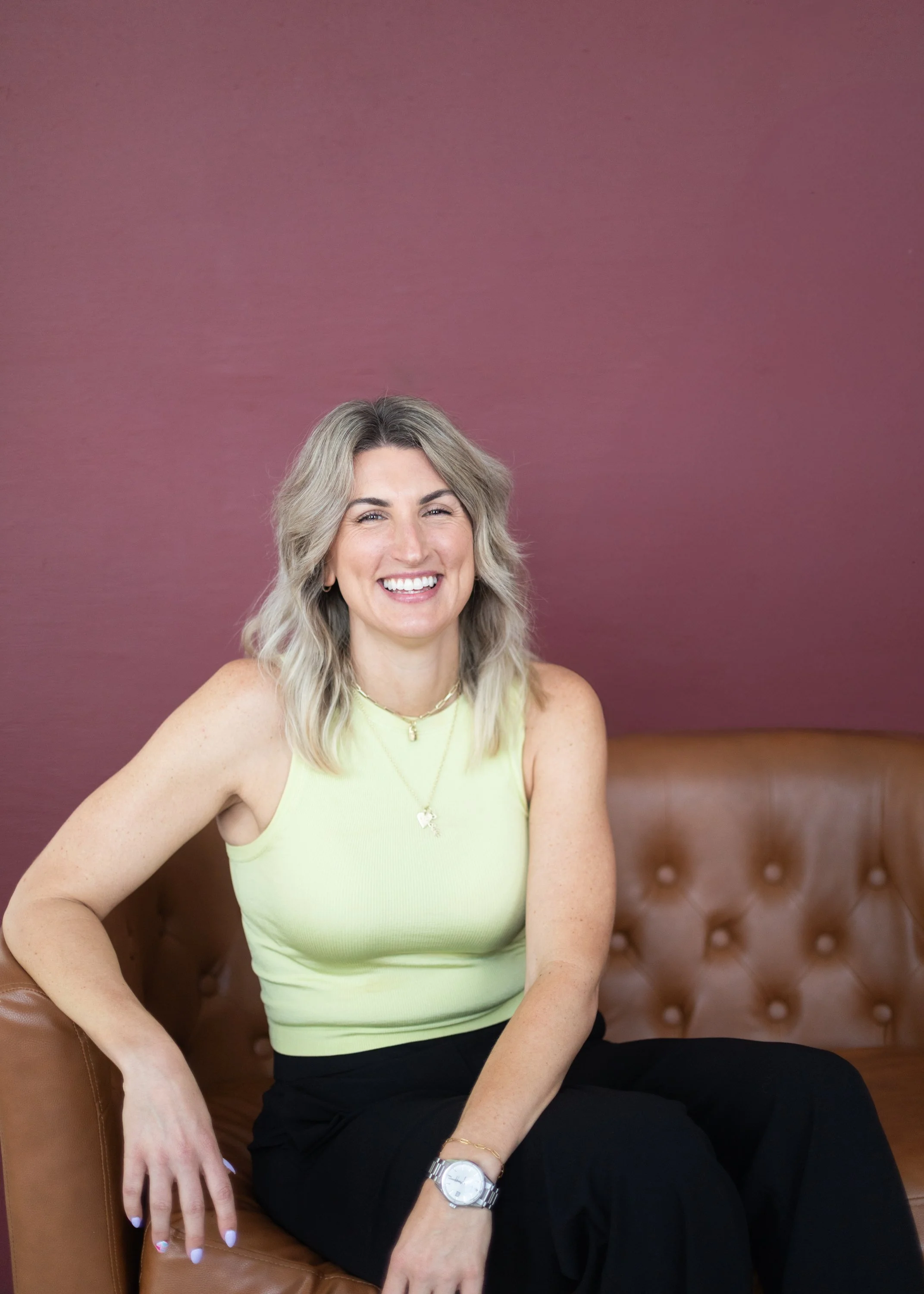 A woman with blonde hair, smiling, wearing a yellow sleeveless top, sitting on a brown leather tufted couch against a purple wall.