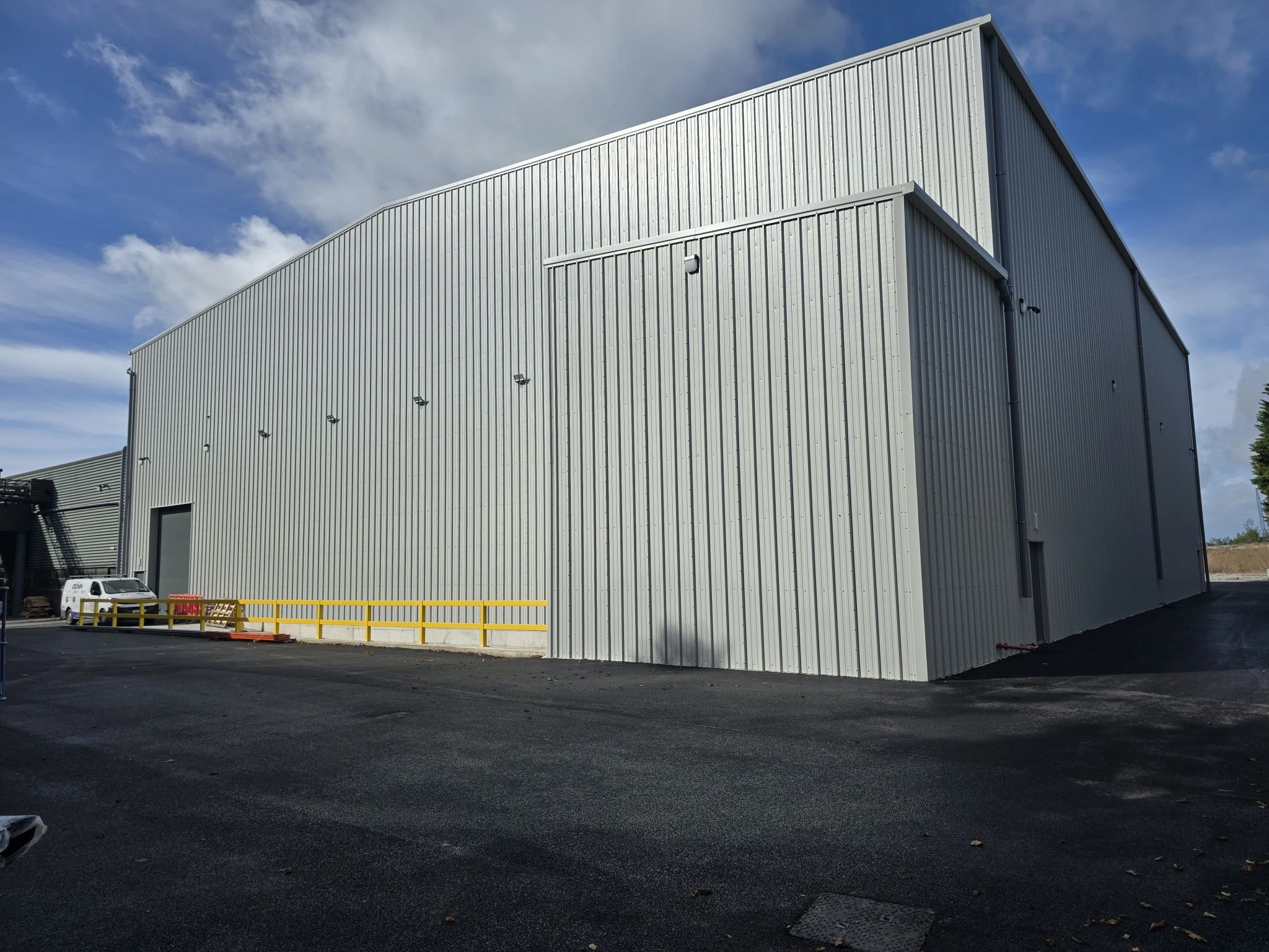 Large white metal industrial building with a roller door, small side door, and a few security cameras. The ground is paved, and there is a truck parked nearby. The sky is partly cloudy.