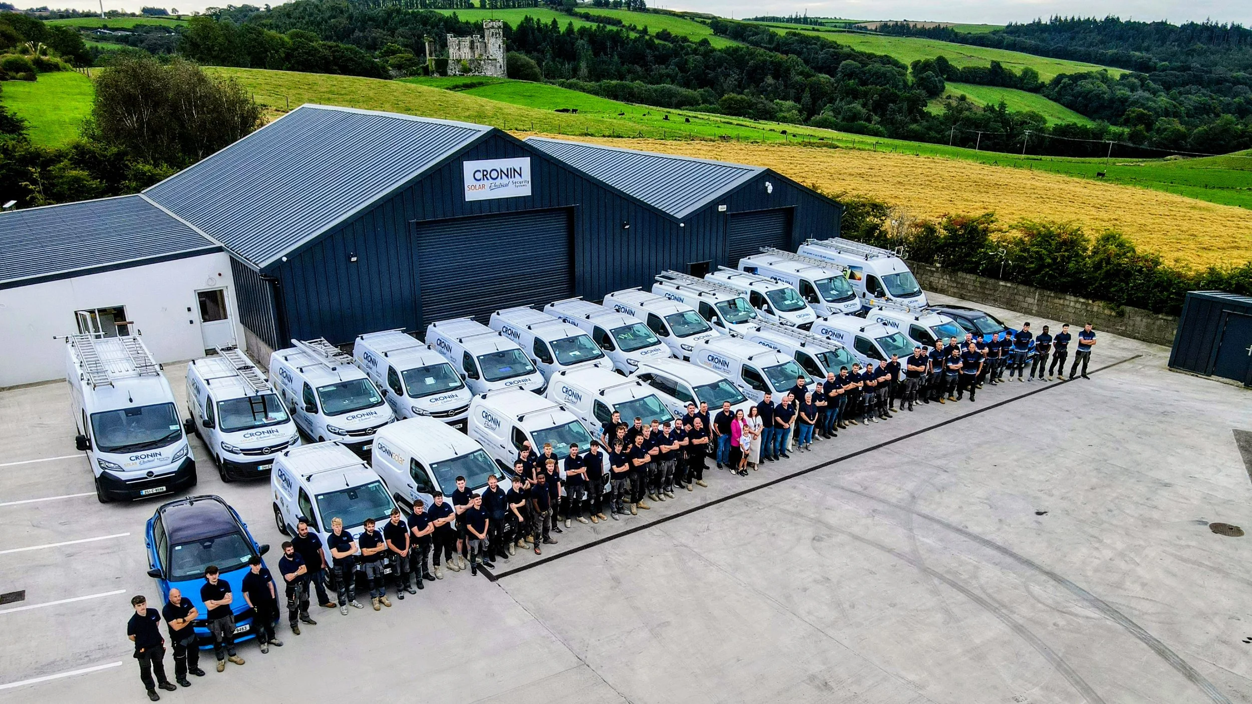 Group of workers standing in front of company vehicles parked outside a building with a sign reading 'CRONIN.' The vehicles are white company vans and one blue car, and the background features a lush green landscape with hills and a castle.