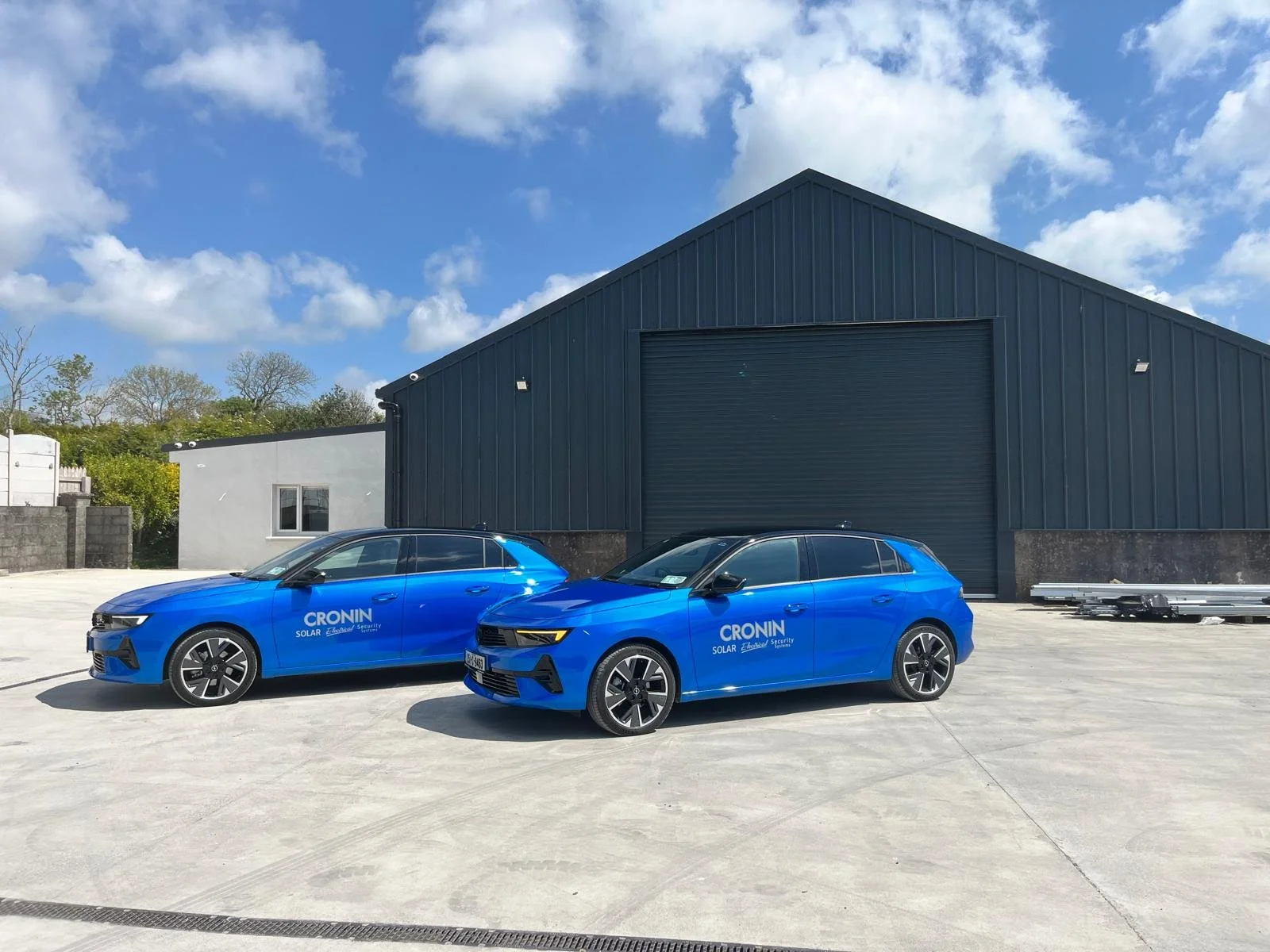 Two blue cars with 'CRONIN' and 'SOLAR' logos parked in front of a black industrial building under a partly cloudy sky.