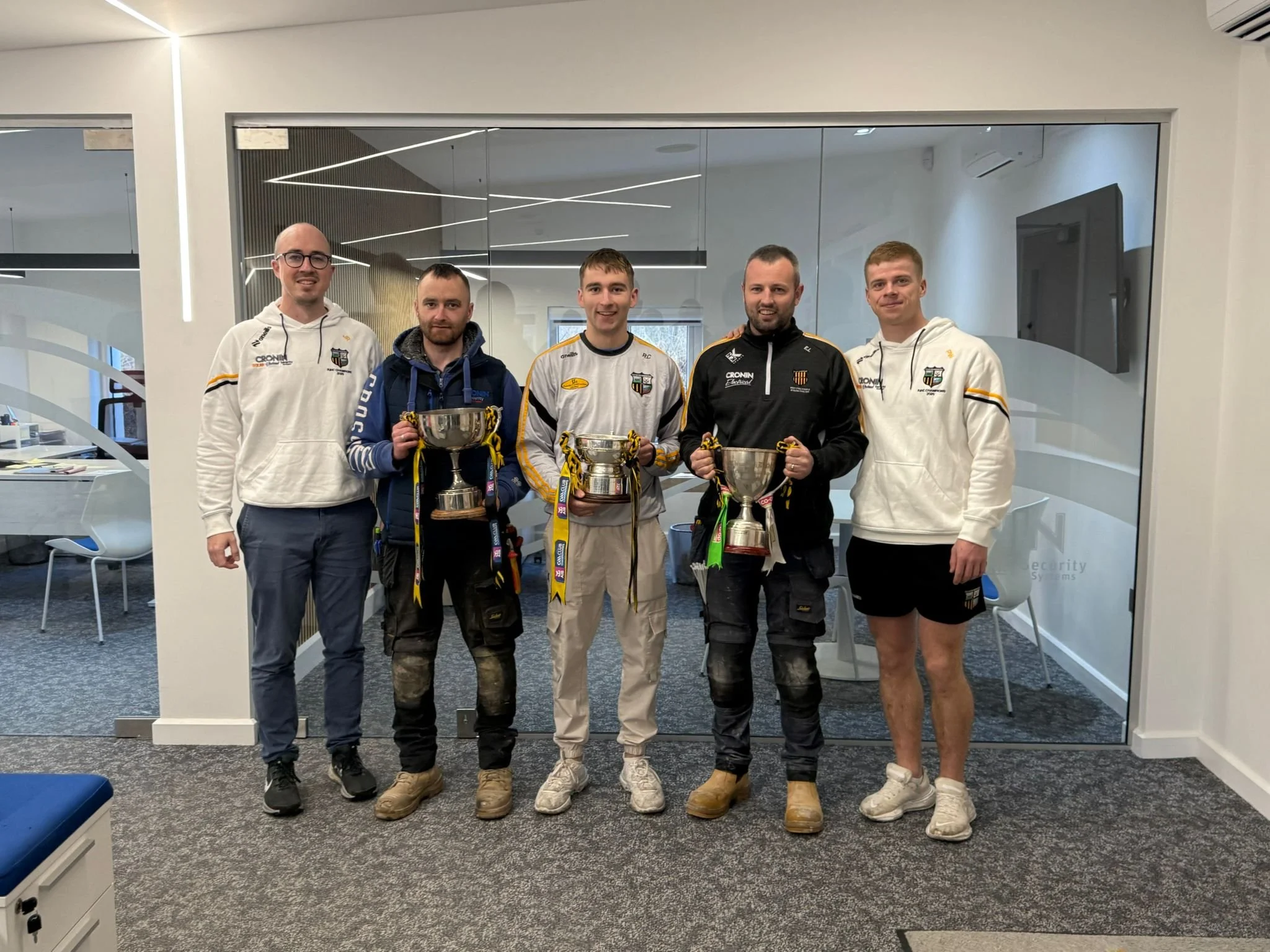 A group of five men standing indoors, holding trophies, with modern lighting and glass walls in the background.