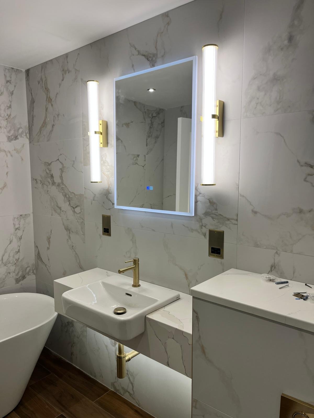 Modern bathroom with marble walls, a rectangular mirror with LED backlight, vertical light fixtures, a white sink with gold fixtures, a white toilet, and a vanity with tools on top.