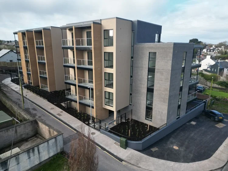 Modern four-story apartment building with balconies, situated on a city street with nearby parking and surrounded by small houses and greenery.