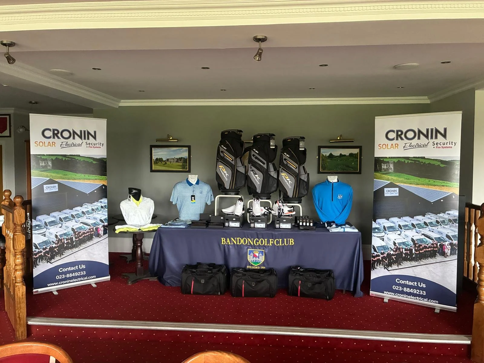 Display of golf equipment and apparel at BanDon Golf Club, including golf bags, shoes, shirts, and training aids, with advertising banners on either side promoting Cronin Solar Electrical Security & Fire Systems.