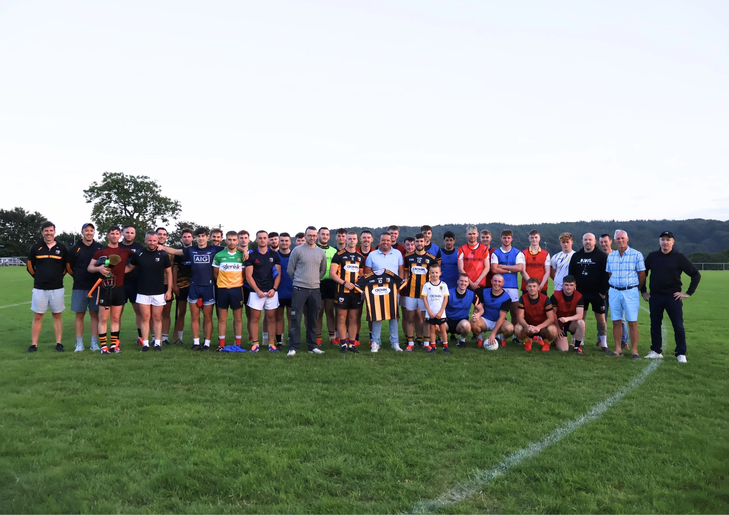 Group of rugby players, coaches, and children on a grassy field, posing for a photo with jerseys and rugby balls, with trees and hills in the background.