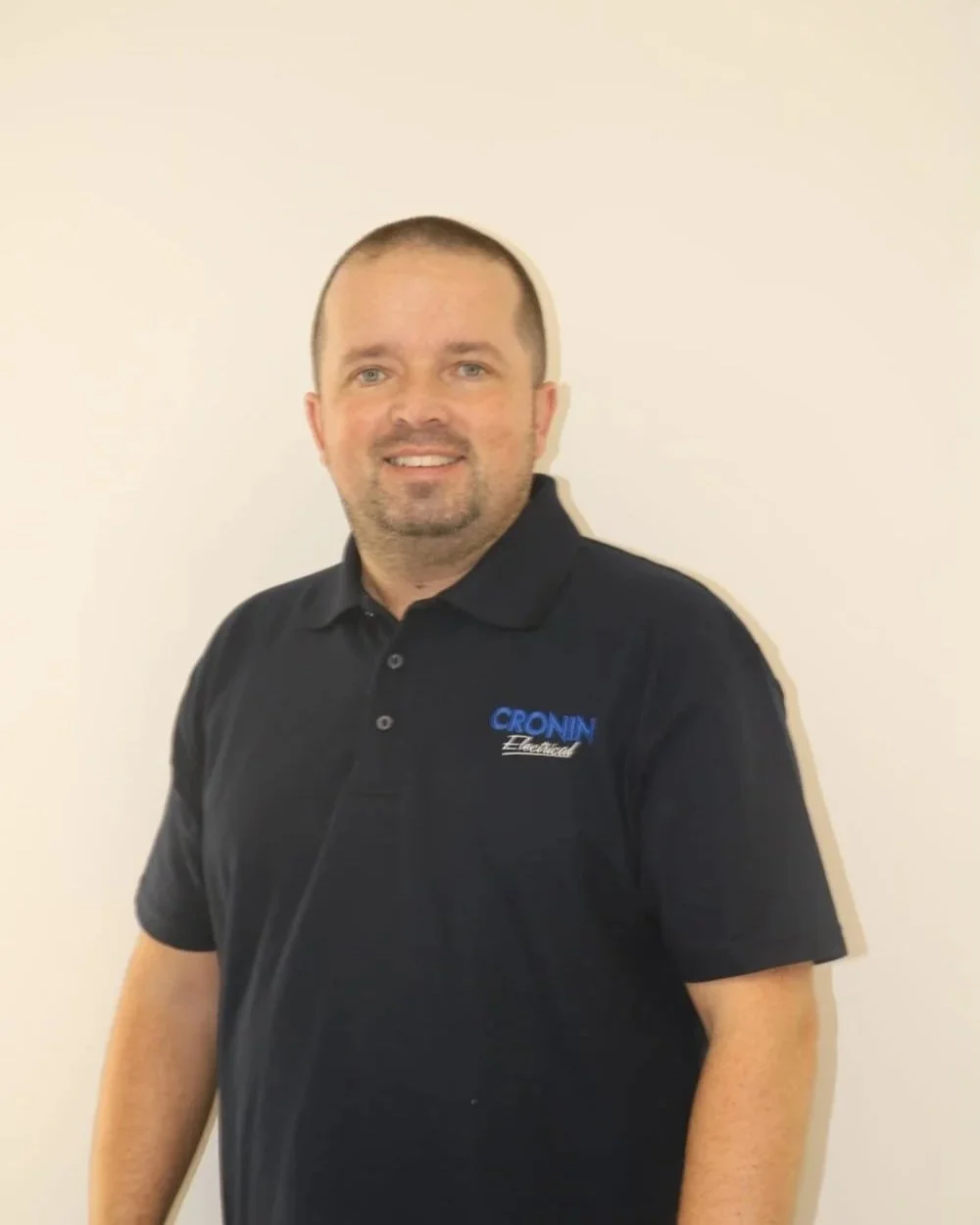 A man with short brown hair and a beard, wearing a black polo shirt with a logo that reads 'CROIN Electrical,' standing against a plain white wall.