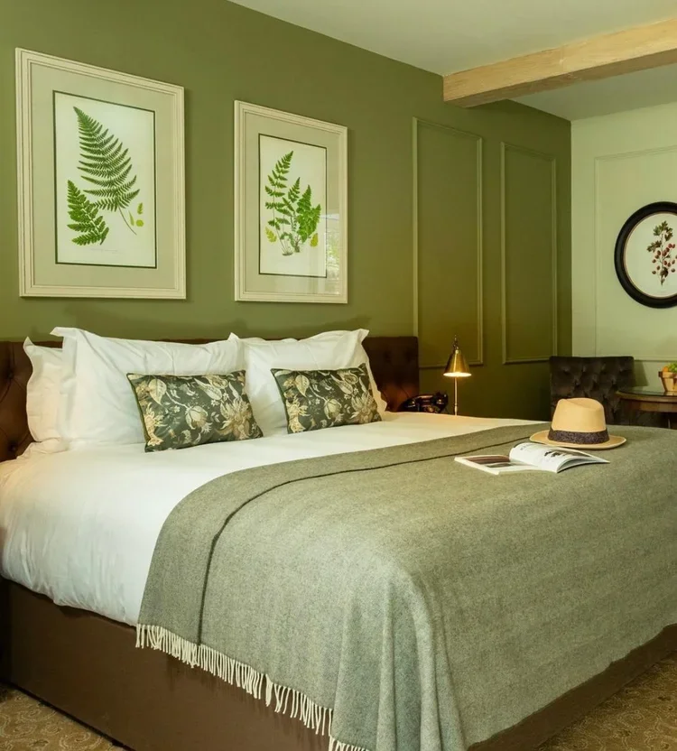 A cozy bedroom with green walls and framed fern prints on the wall. The bed has white pillows, floral decorative pillows, a gray blanket, and a straw hat on top. There is a small night lamp, a book, and a telephone on a side table. There are additional chairs and artwork in the background.