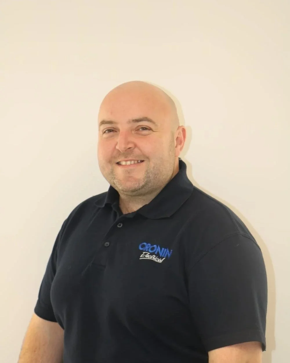 A smiling man with a shaved head wearing a black polo shirt with 'Cronin Electrical' embroidered on it, standing against a plain off-white wall.