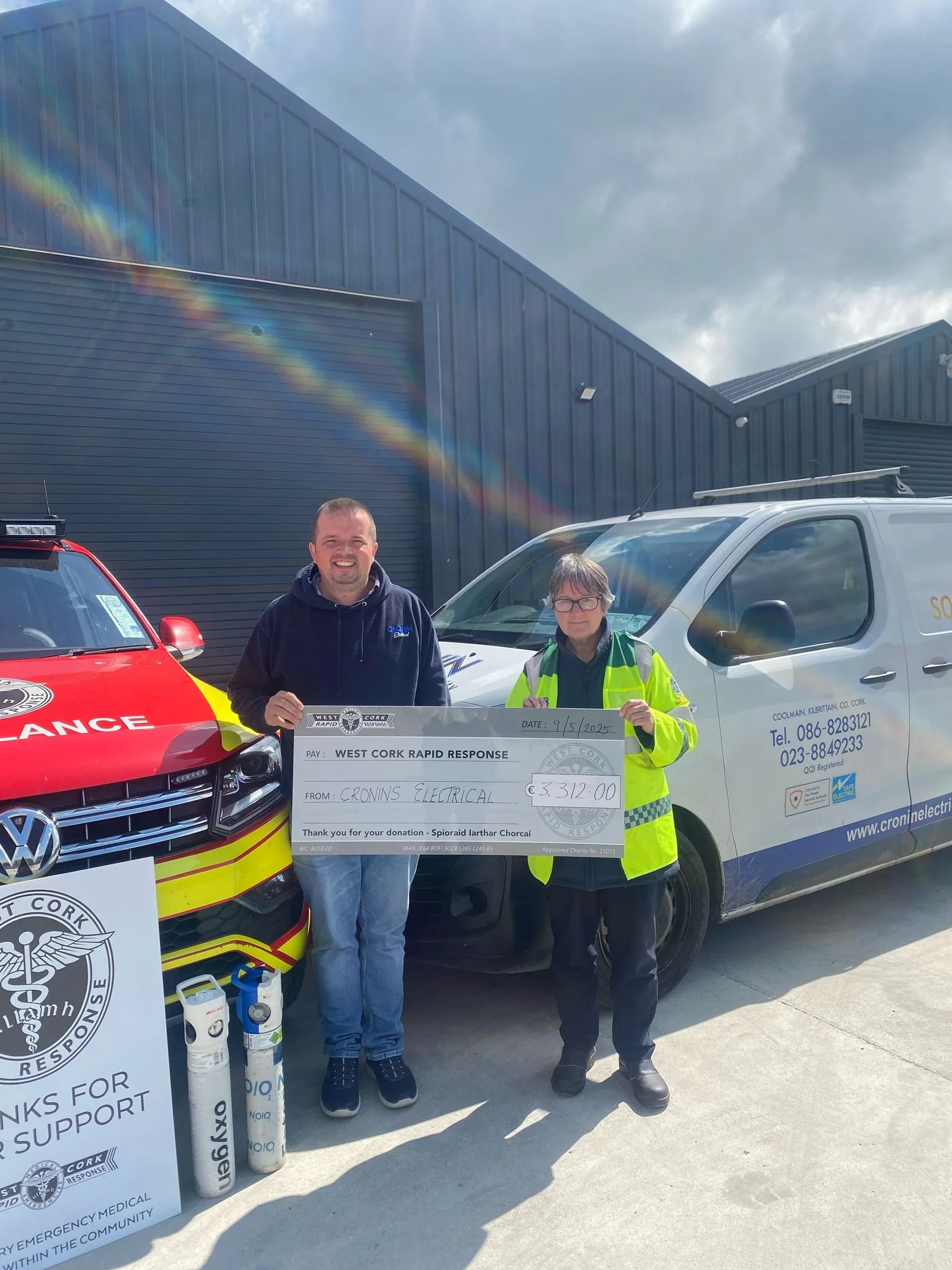 Two men standing in front of a white van and a police car holding a large donation check for €3312.00 made out to West Cork Rapid Response, date 9/5/2025. One man is dressed in a dark hoodie and jeans, and the other in a high-visibility jacket with glasses. There are signs and bottles on the ground supporting the donation.