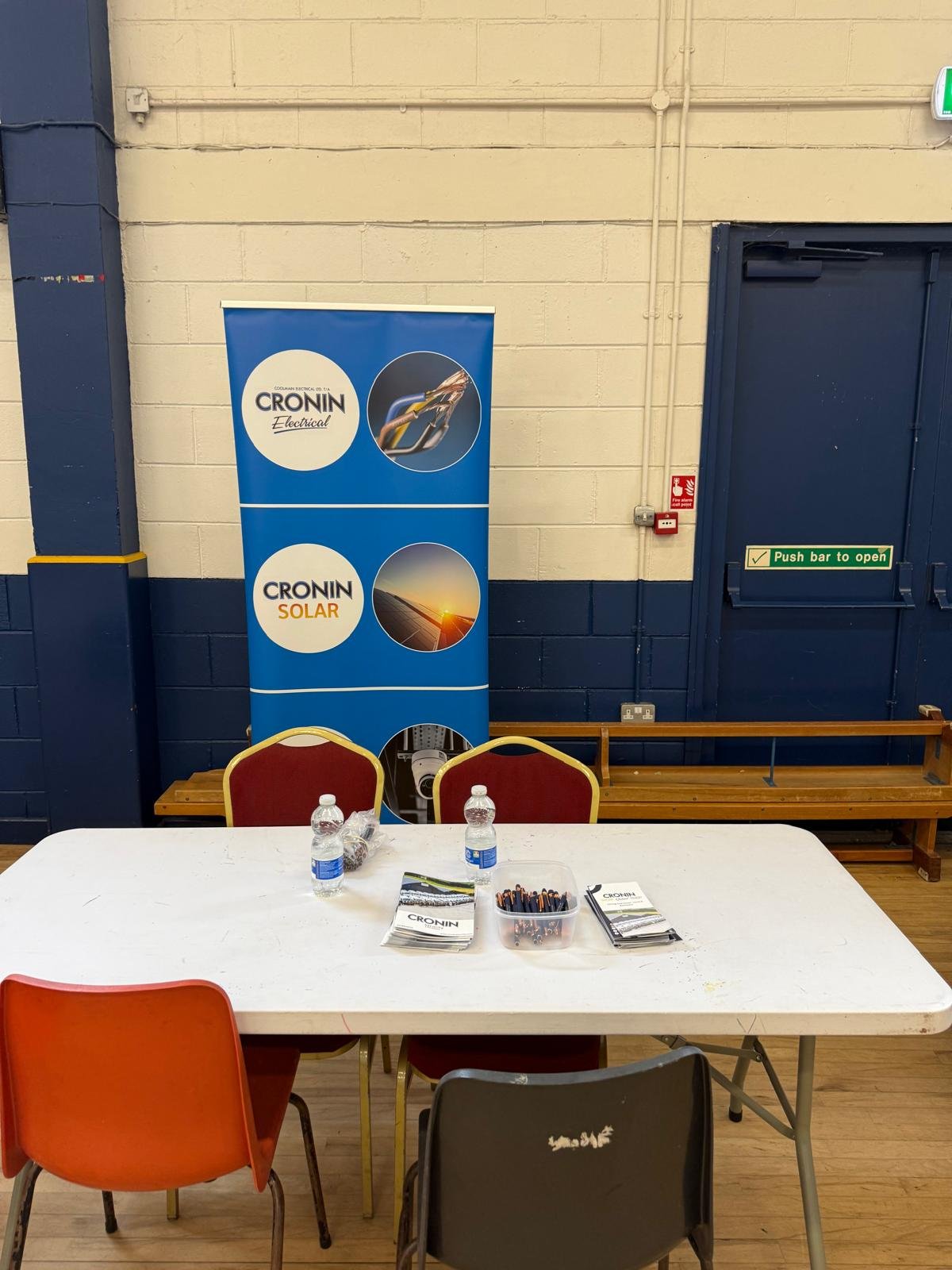 Indoor meeting area with a white table, three chairs, water bottles, brochures, and a plastic container with pens. Behind the table is a blue banner with white circles and text advertising Cronin Electrical and Solar services. To the right is a door 