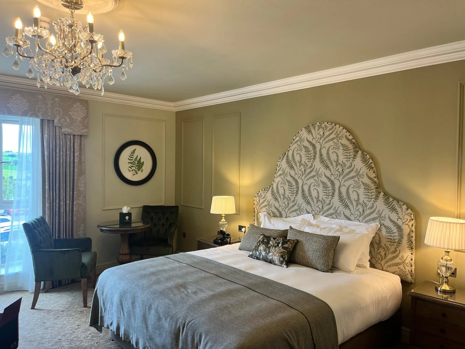 A luxurious hotel room with a large bed, ornate headboard, and two bedside lamps. There are two chairs and a small table near a window adorned with drapes, and a chandelier hanging from the ceiling.