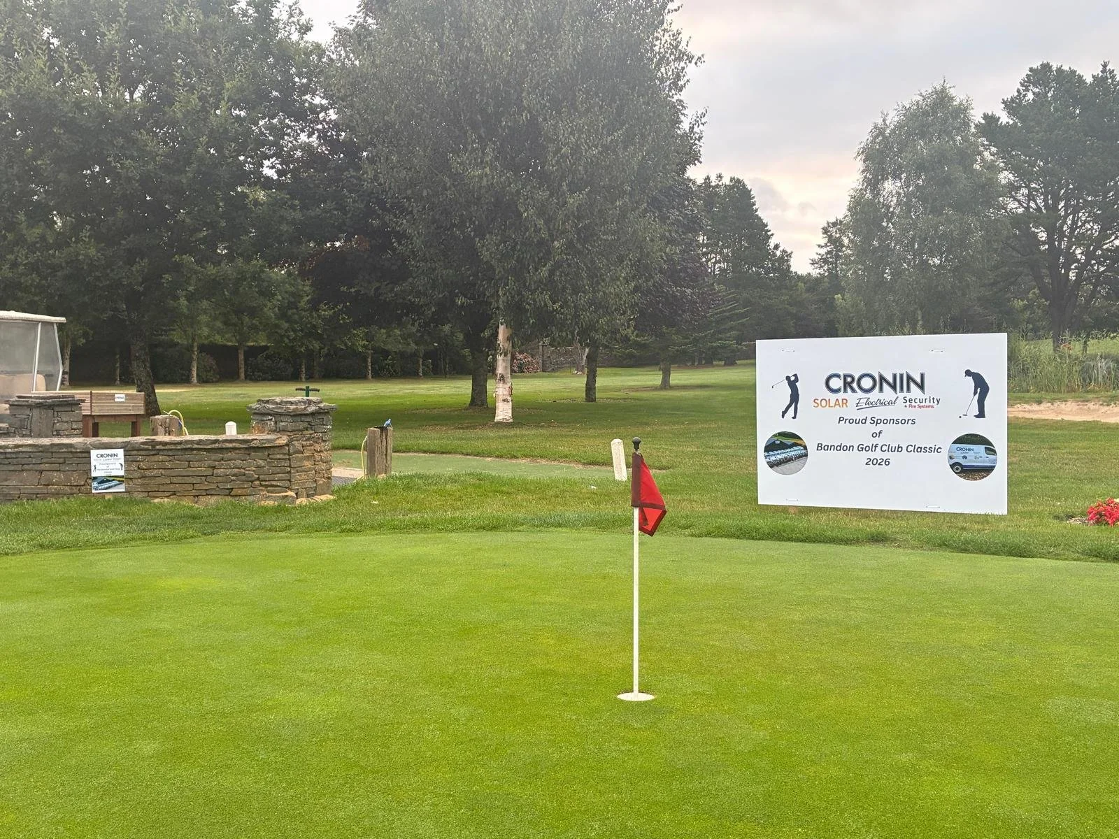 A golf course with a putting green in the foreground and a sand trap to the right. There is a large advertising banner on the right side promoting Cronin Solar, Electrical & Security for the Bandon Golf Club Classic 2026. The background features trees and a cloudy sky.