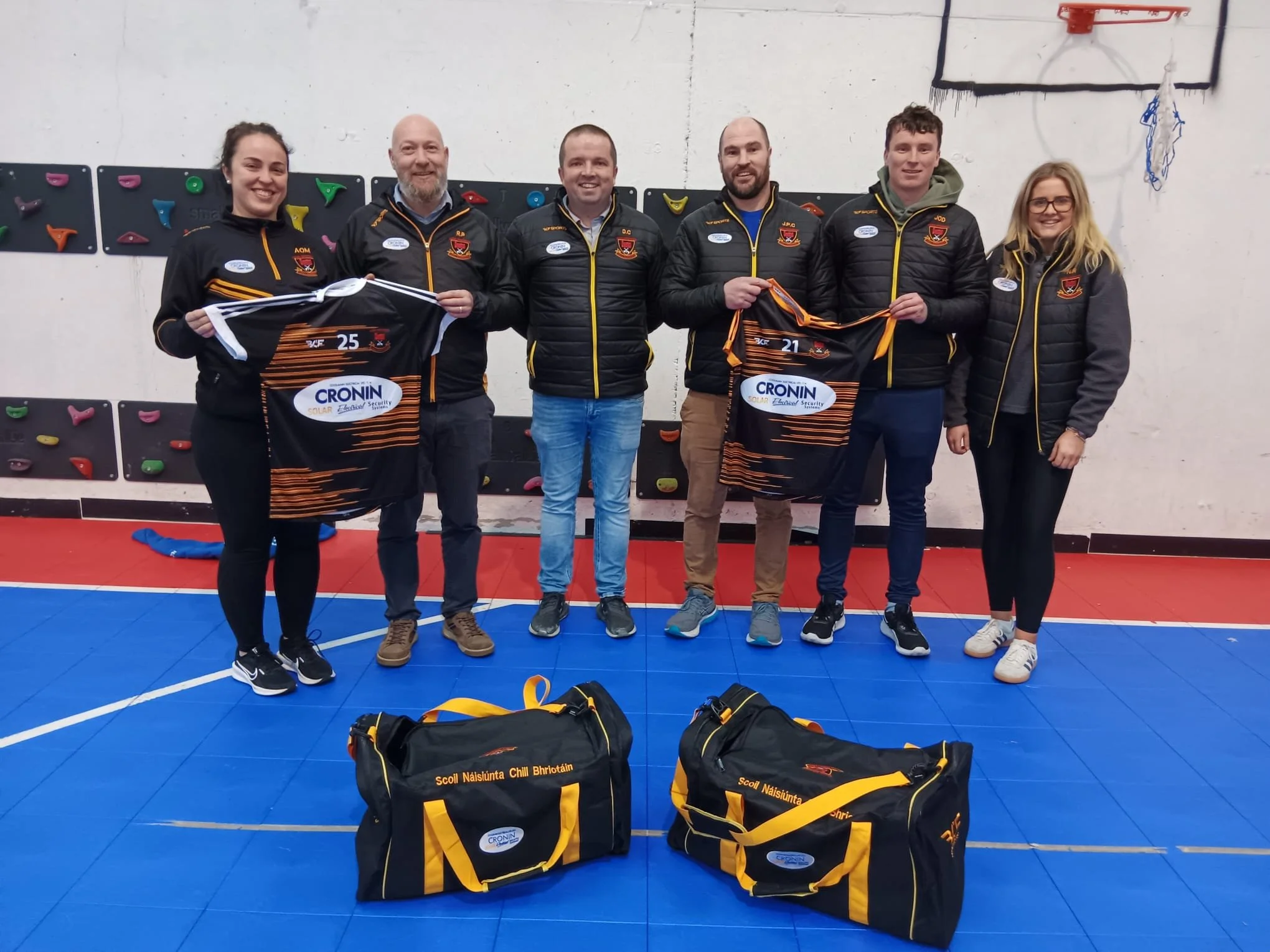 Six people standing indoors on a sports court, holding sports jerseys, smiling for the camera. The background features climbing holds on a wall and a basketball hoop. There are bags and sports equipment on the floor in front of them.