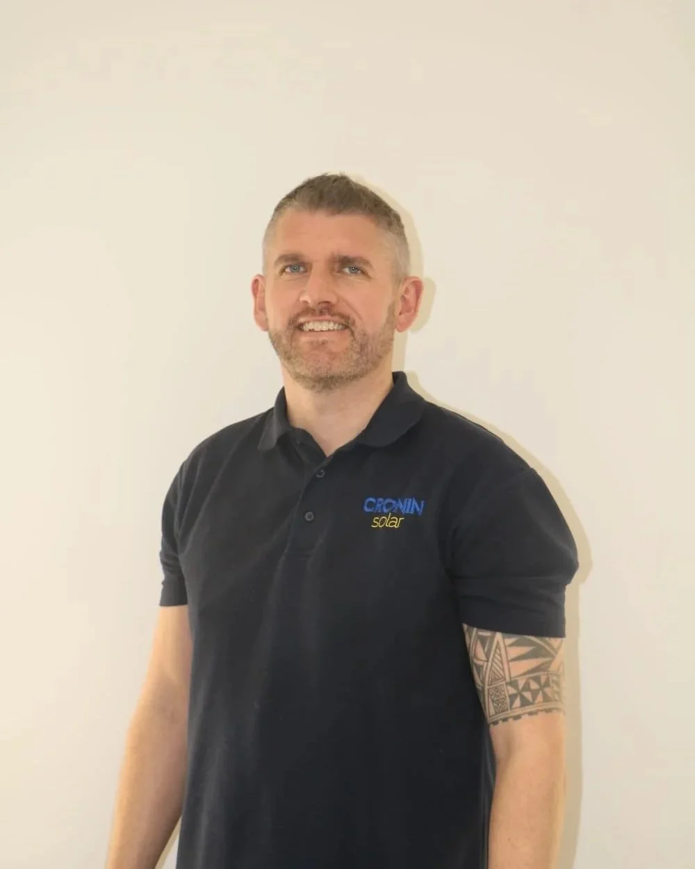 A man with short light brown hair and a beard, wearing a black polo shirt with a logo that says 'CRONIN solar' on it, standing in front of a plain white wall.