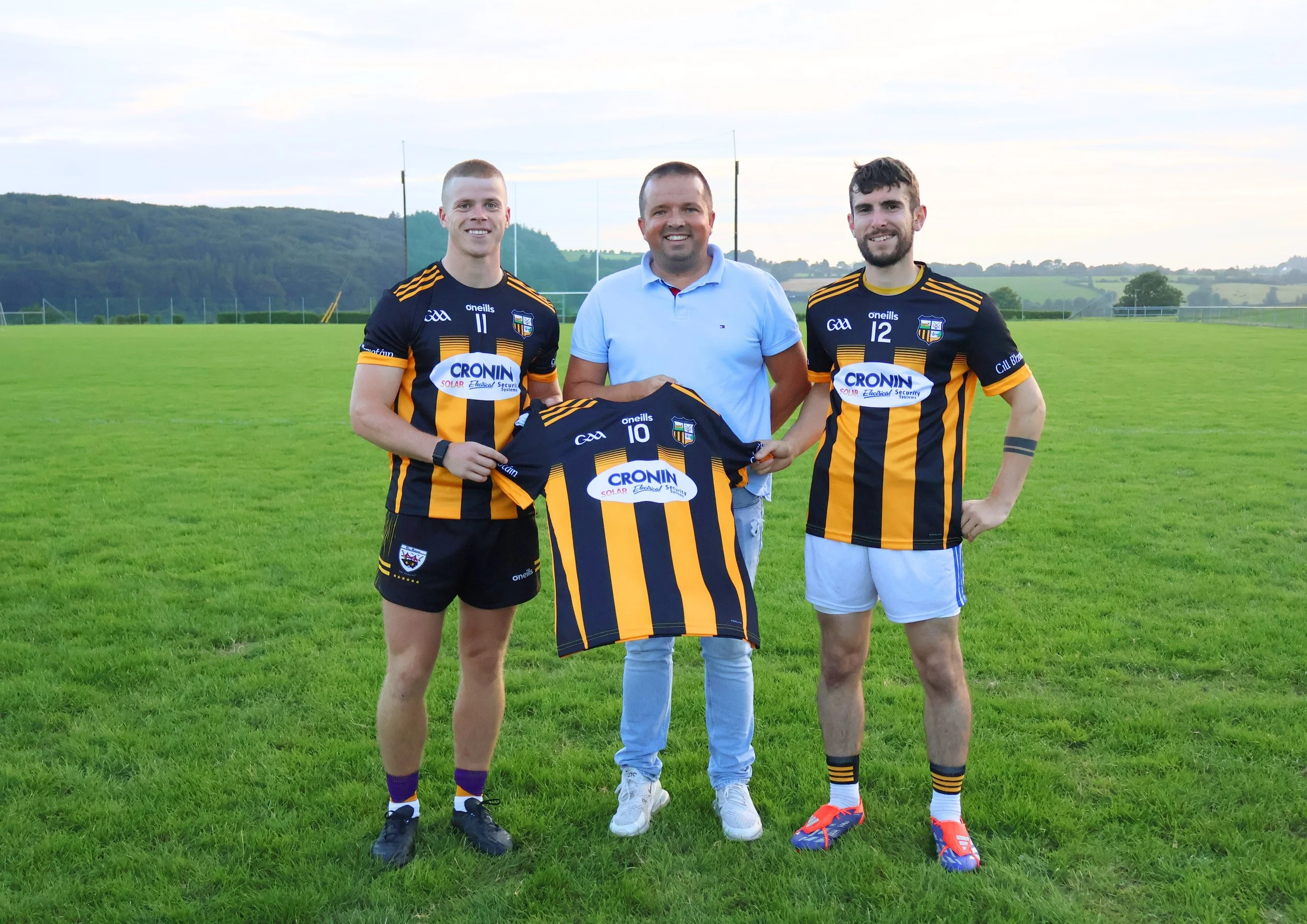 Three men standing in a grassy field, two are wearing black and yellow striped soccer jerseys, one in a light blue shirt holding a soccer jersey, with a scenic background of hills and trees.