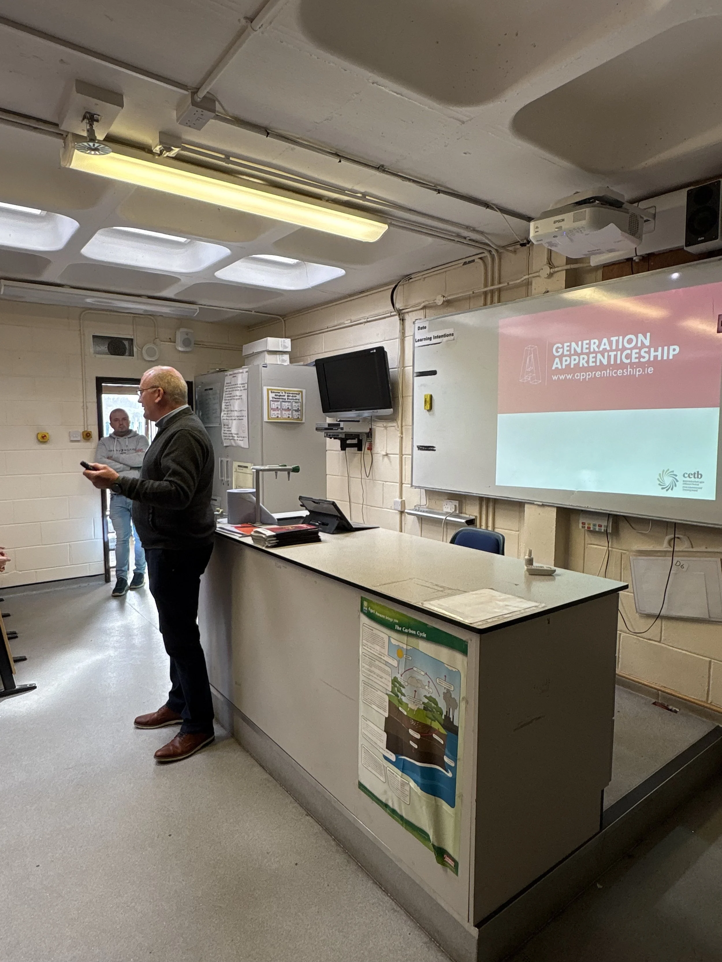 A classroom with a man standing at a counter, a whiteboard with a presentation slide, and a person entering through a door in the background.