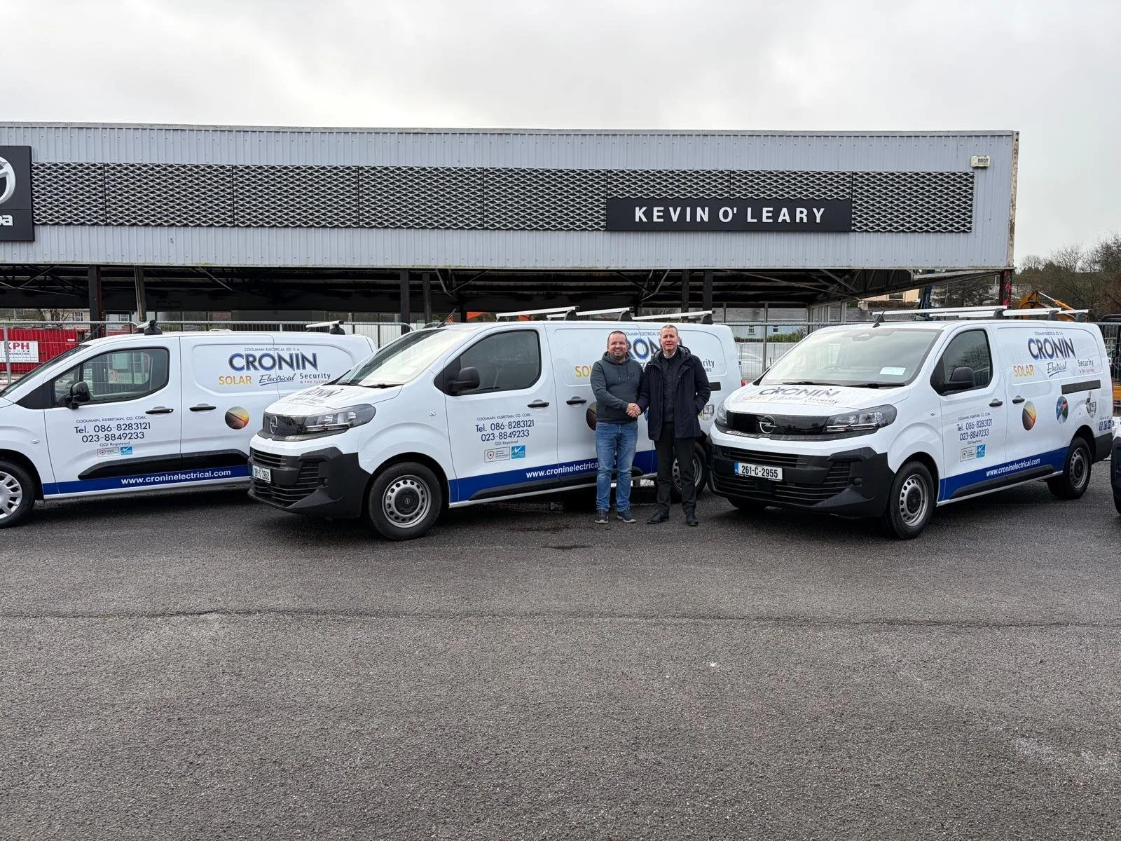 Two men standing between two white company vans with company logos, parked in front of a building with a sign reading 'KEVIN O'LEARY'.
