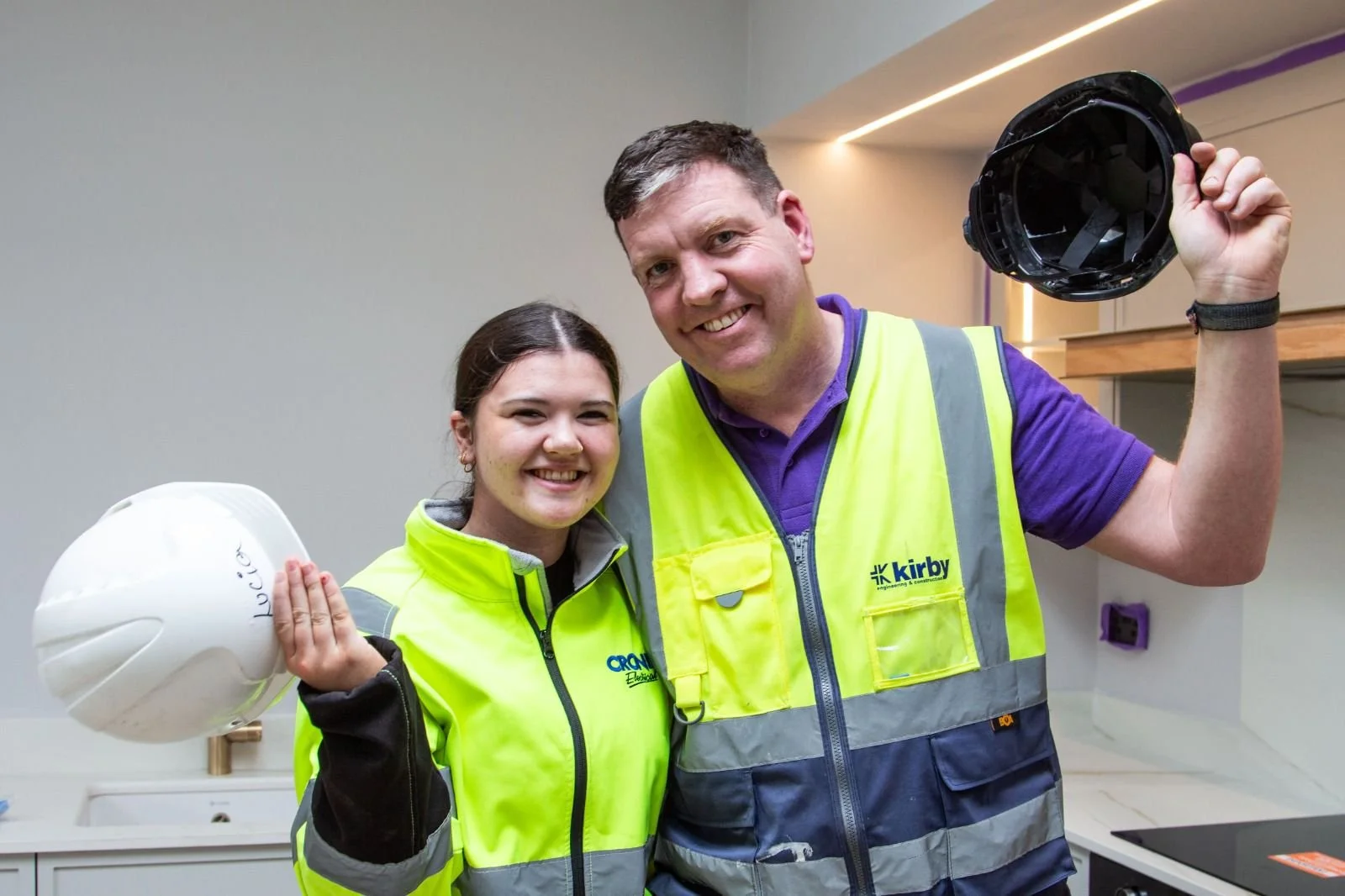 Two construction workers smiling and holding hard hats, standing indoors with a modern kitchen background.