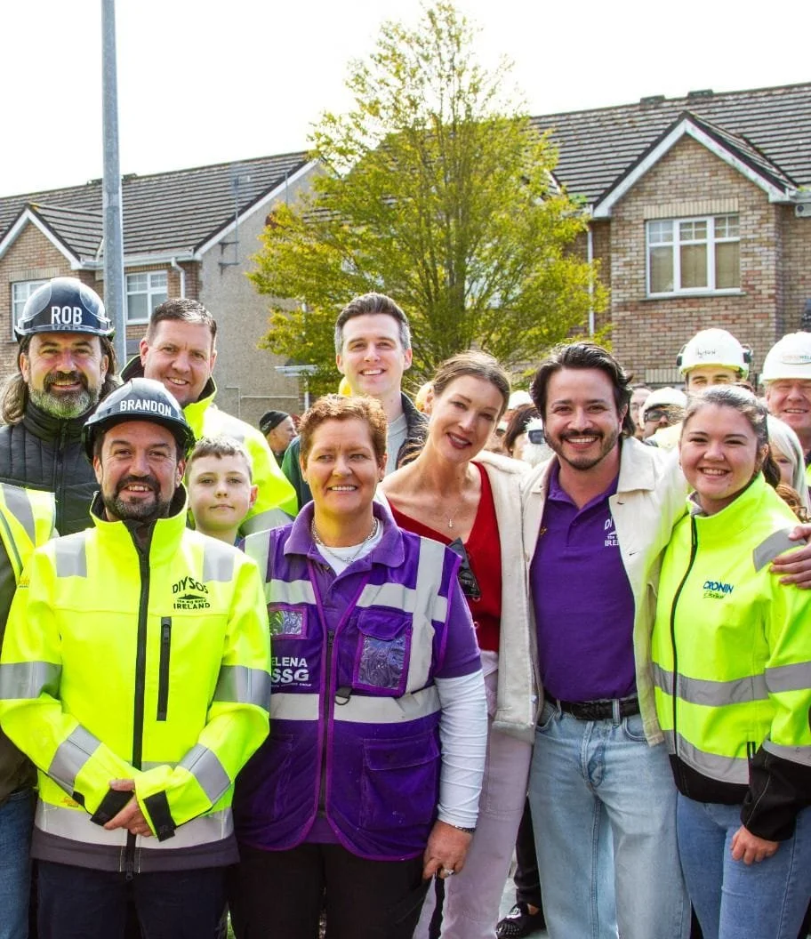 Group of people gathered outdoors, some wearing safety helmets and high-visibility jackets, with houses and a tree in the background.