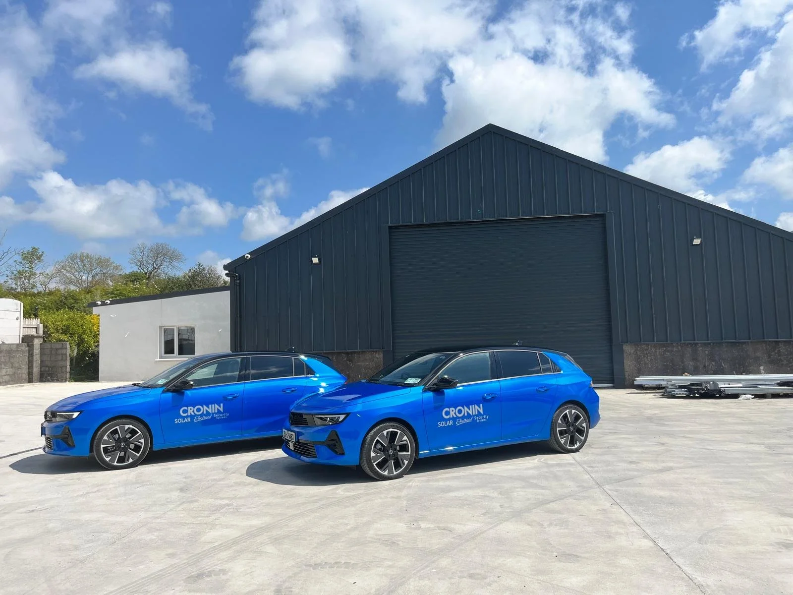 Two blue electric vehicles parked in front of a large industrial building with a black garage door, under a partly cloudy sky.