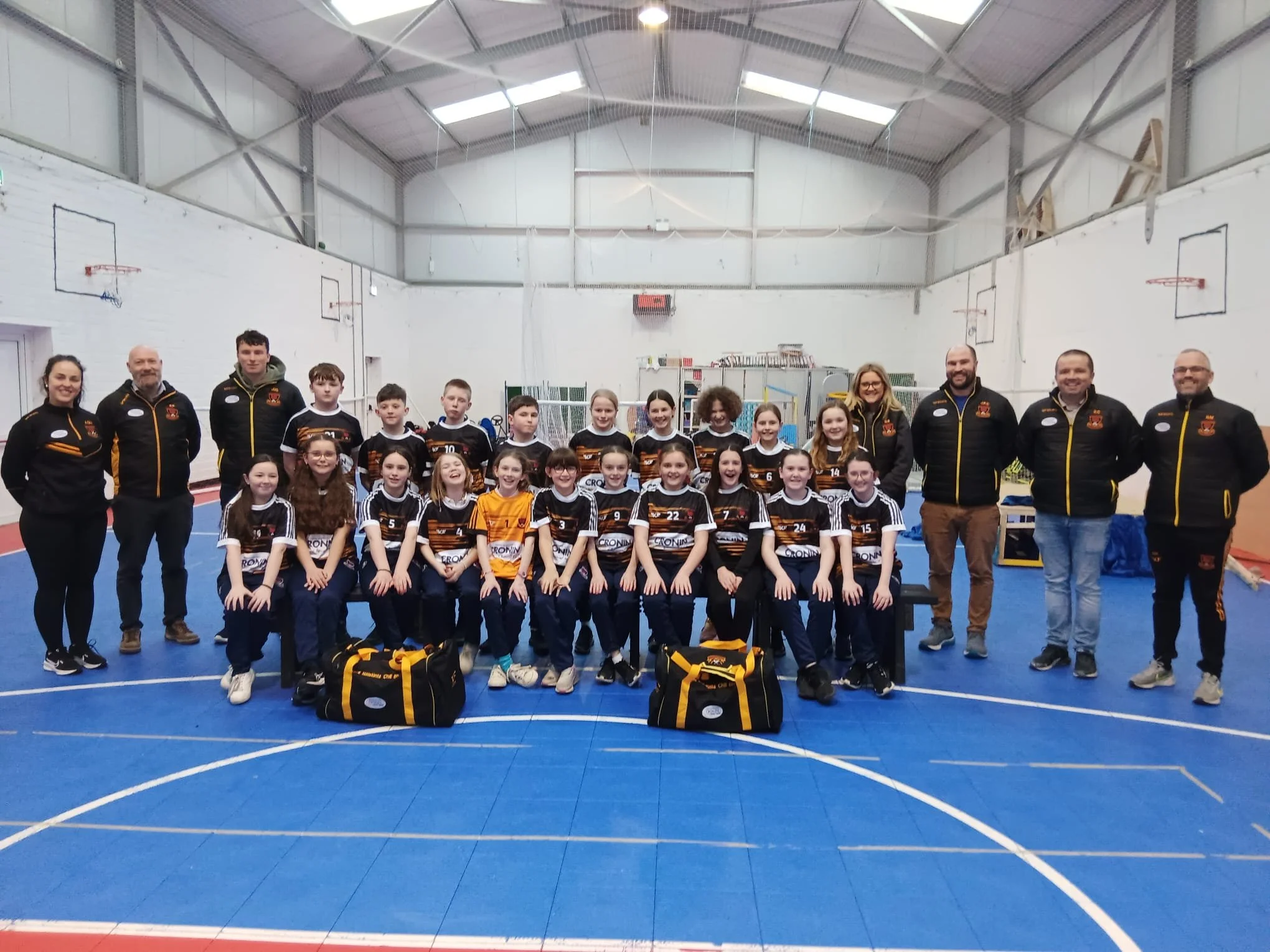 A youth volleyball team and their coaches posing for a group photo in an indoor gymnasium with basketball hoops, some children seated in front and others standing behind, all wearing team uniforms and surrounded by sports bags.
