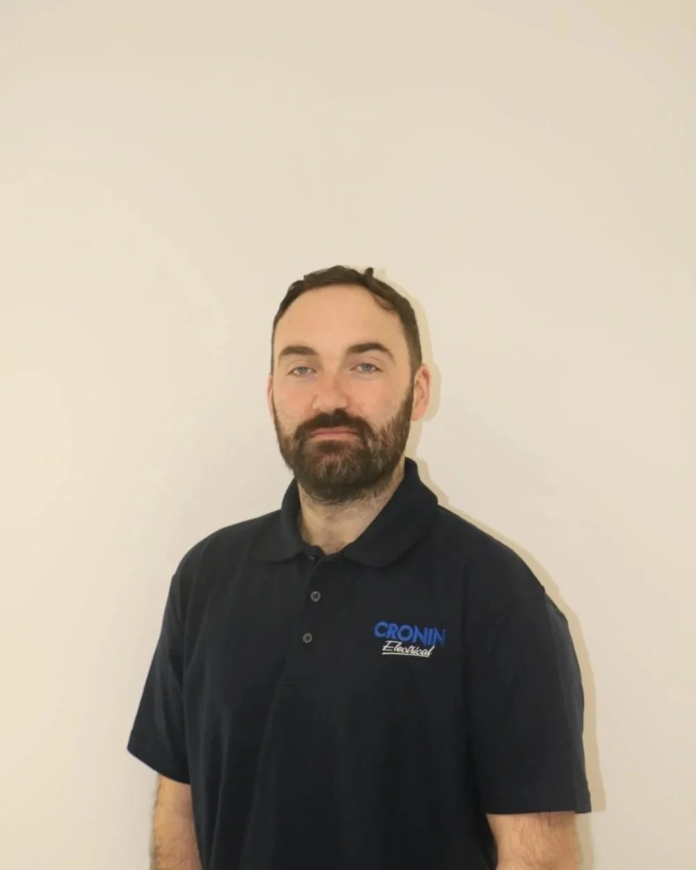 A man with a beard wearing a black polo shirt with the logo 'CRONIN Electrical' stands against a plain beige wall.