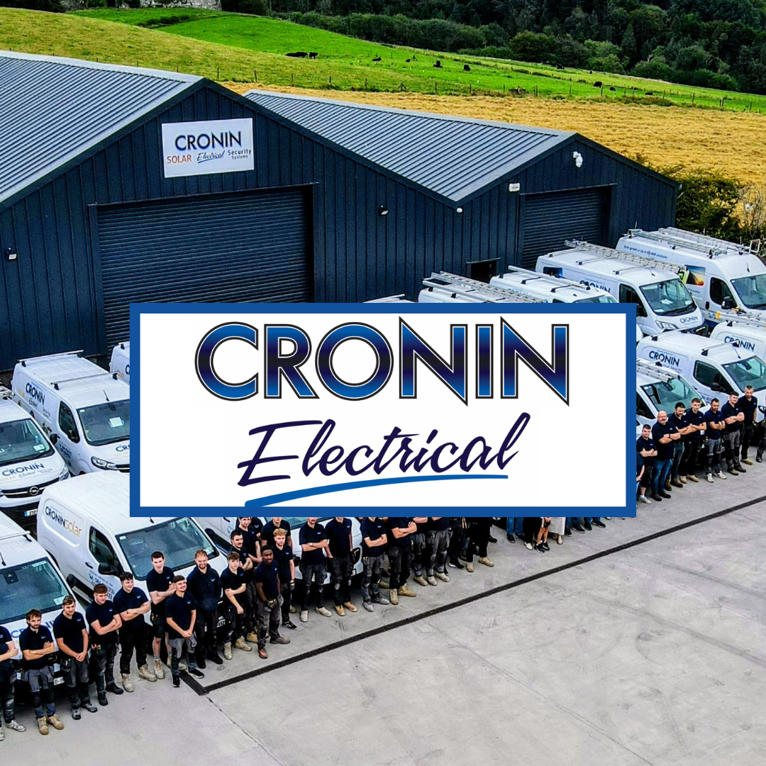 A fleet of white service vans with the company logo parked in front of a large blue industrial building with a sign that reads 'CRONIN Solar Electrical Security.' A group of employees in matching navy shirts stands in front of the vans. Green fields and trees are visible in the background.