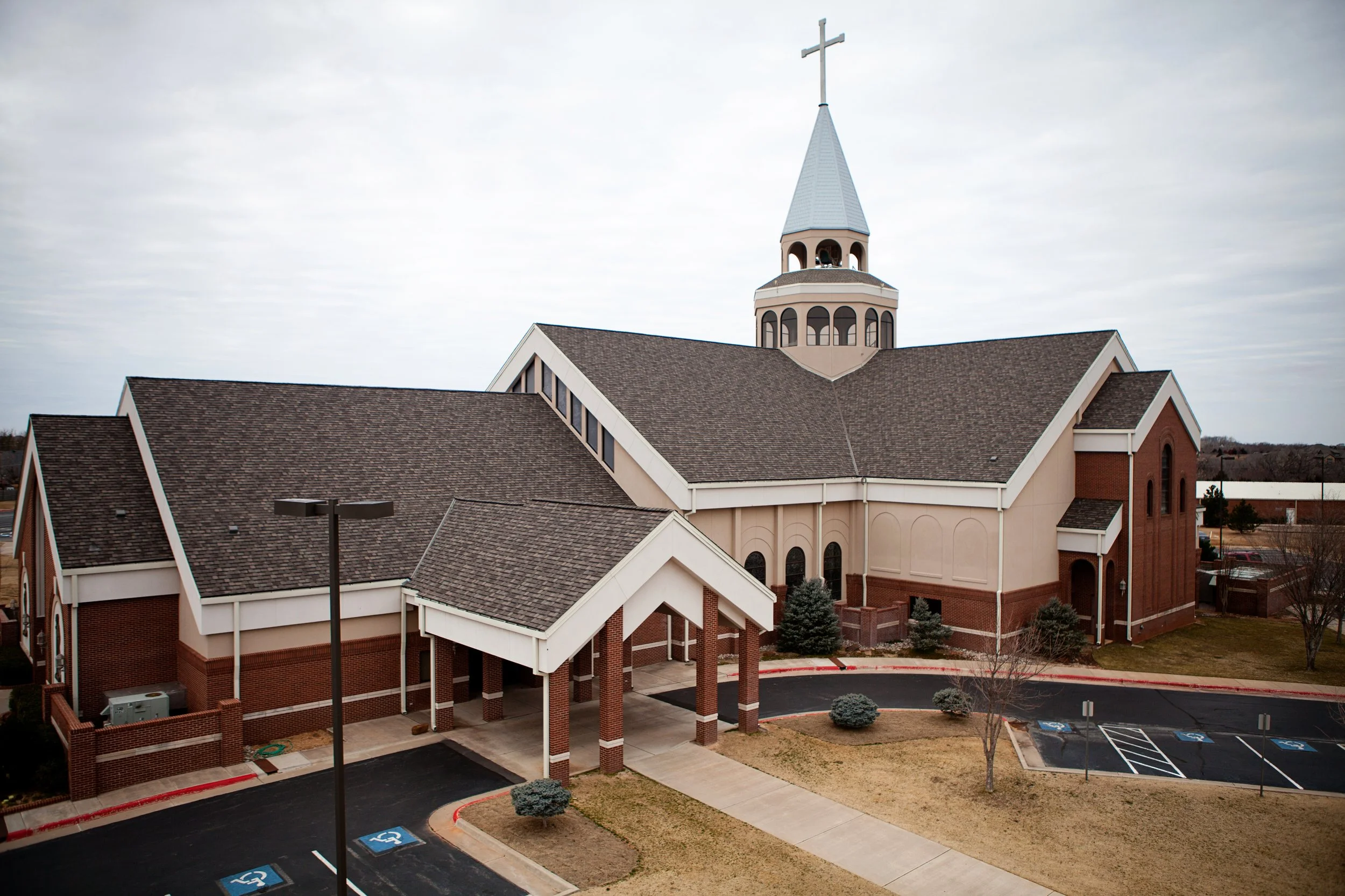 A church building with a tall steeple and cross on top, surrounded by small landscaped trees, parking spaces including designated handicapped spots, and a sidewalk in front.