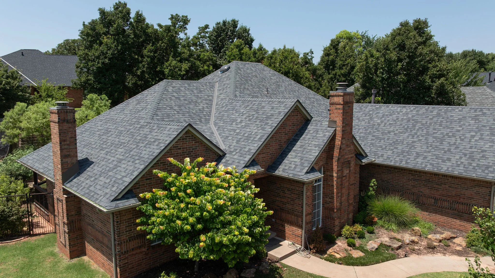 A suburban house with red brick walls and a gray shingle roof, surrounded by lush green trees and landscaping, under a clear blue sky.