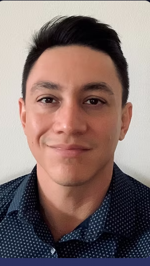 A young man with short, dark hair and light skin, smiling slightly, wearing a dark blue, patterned button-up shirt, standing against a white wall.