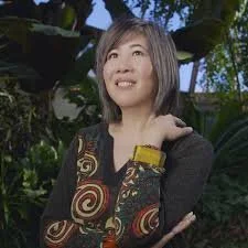 A young woman with shoulder-length hair and a patterned shirt, standing outdoors in front of lush green plants, looking up with a smile.
